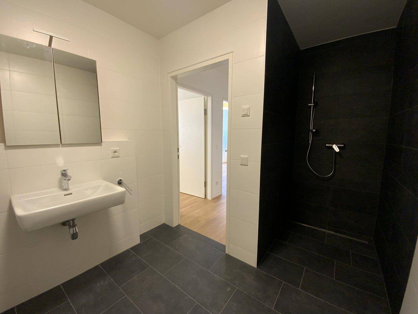Zeitgemäßes Badezimmer mit ebenerdiger Dusche, Waschbecken und dunklen Bodenfliesen.
