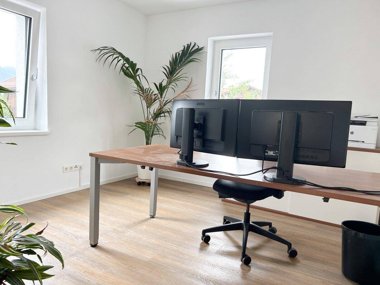 Helles Büro mit zwei Monitoren, Schreibtisch und großen Fenstern, die viel Tageslicht hereinlassen.