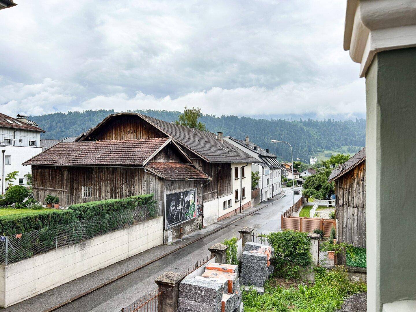 Blick auf die Straße und umliegende Gebäude mit bewaldeten Hügeln im Hintergrund unter bewölktem Himmel.