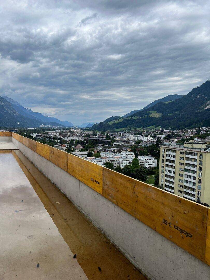 Panoramablick von einer Baustelle auf eine Stadt und umliegende Berge unter einem bewölkten Himmel.