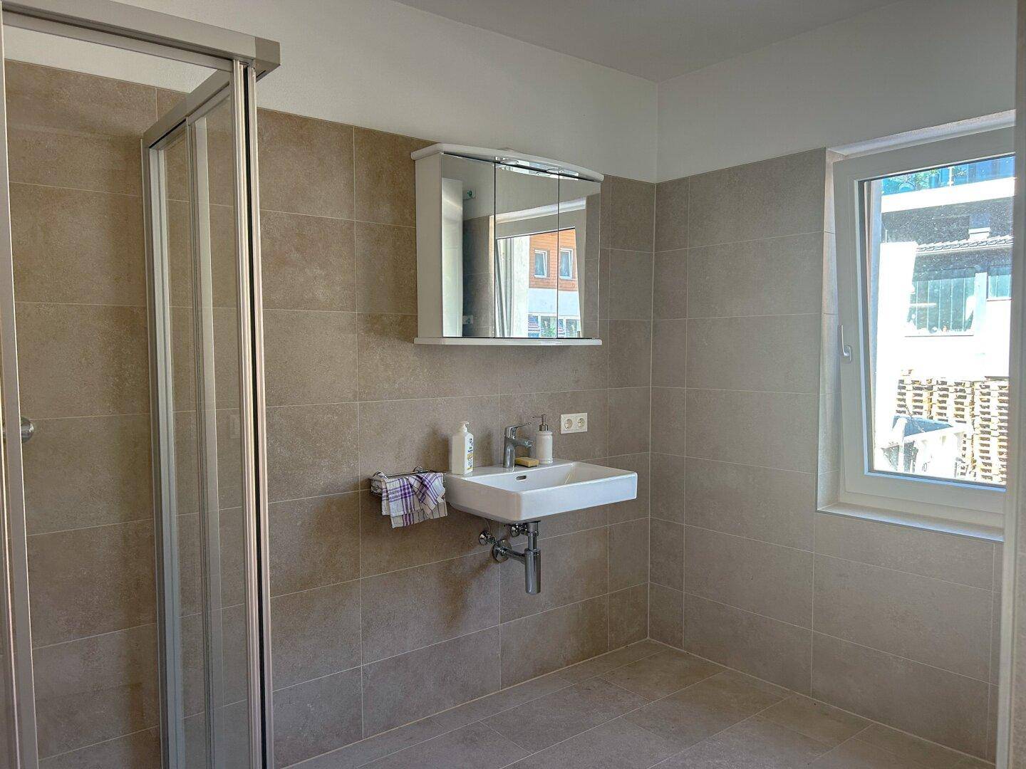 Modernes Badezimmer mit beigefarbenen Fliesen, Duschkabine und einem Fenster für Tageslicht.