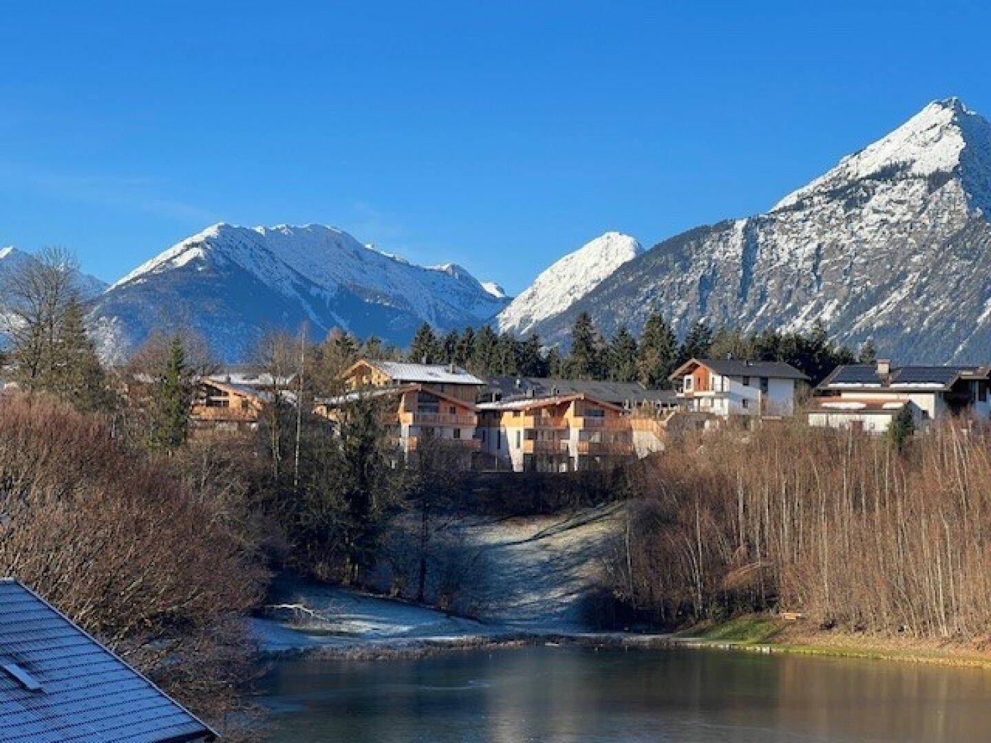 Malerische Winterlandschaft mit einem See, Häusern und schneebedeckten Bergen unter blauem Himmel.