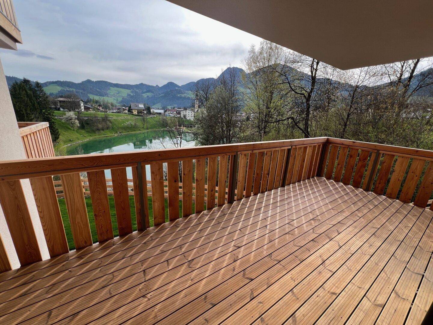 Geräumiger Holzbalkon mit sonnigem Blick auf einen See und die umliegende Berglandschaft.
