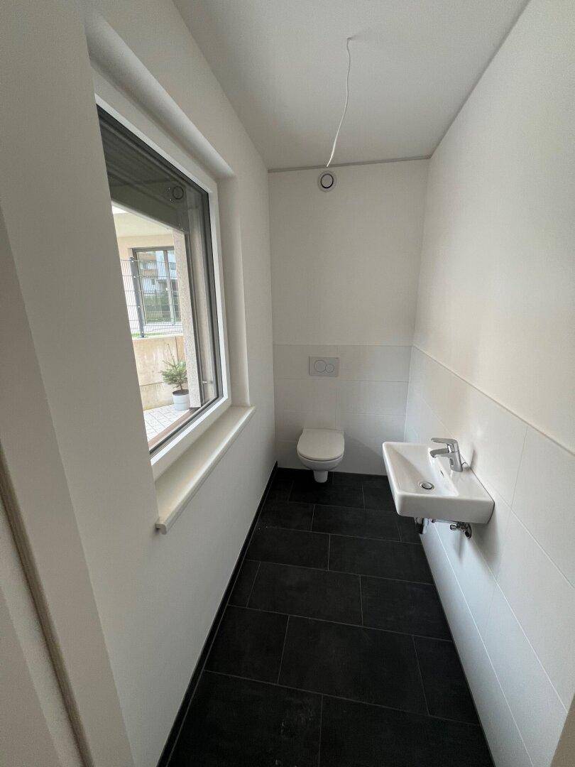 Gäste-WC mit Fenster, wandhängender Toilette und kleinem Waschbecken auf dunklem Fliesenboden.