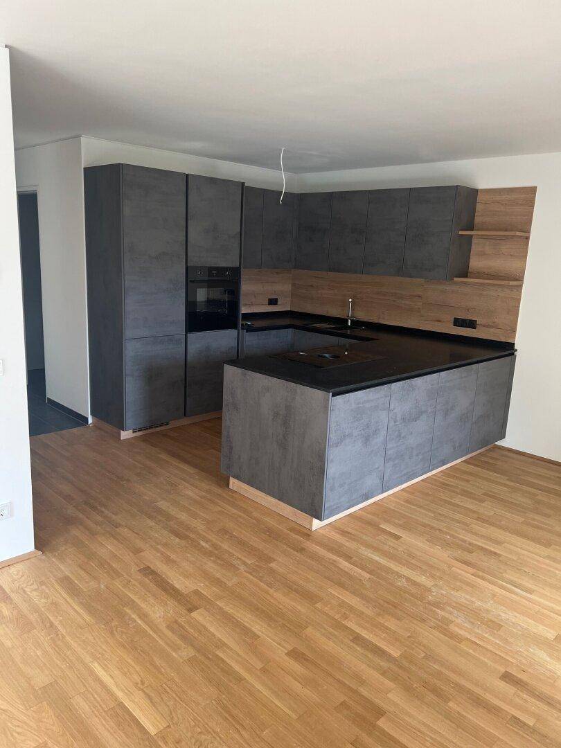 Moderne Küche mit grauen Schränken, schwarzer Arbeitsplatte und Kochinsel auf hellem Holzboden.