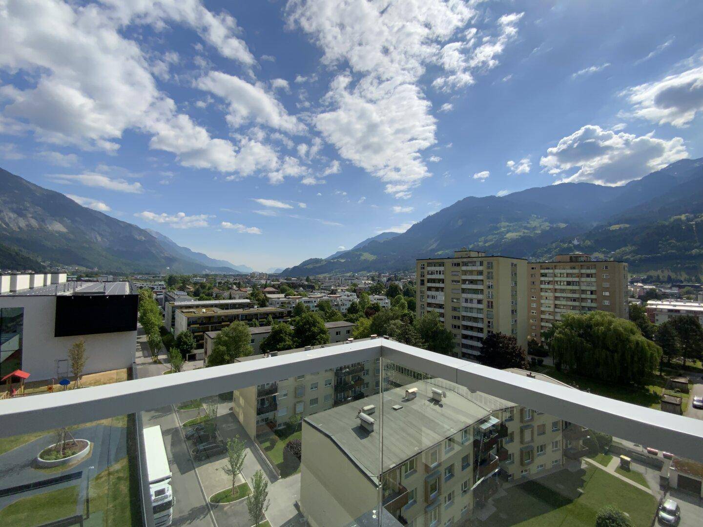 Balkon mit Glasbrüstung und weitem Blick über die Stadt und die majestätischen Berge unter blauem Himmel.