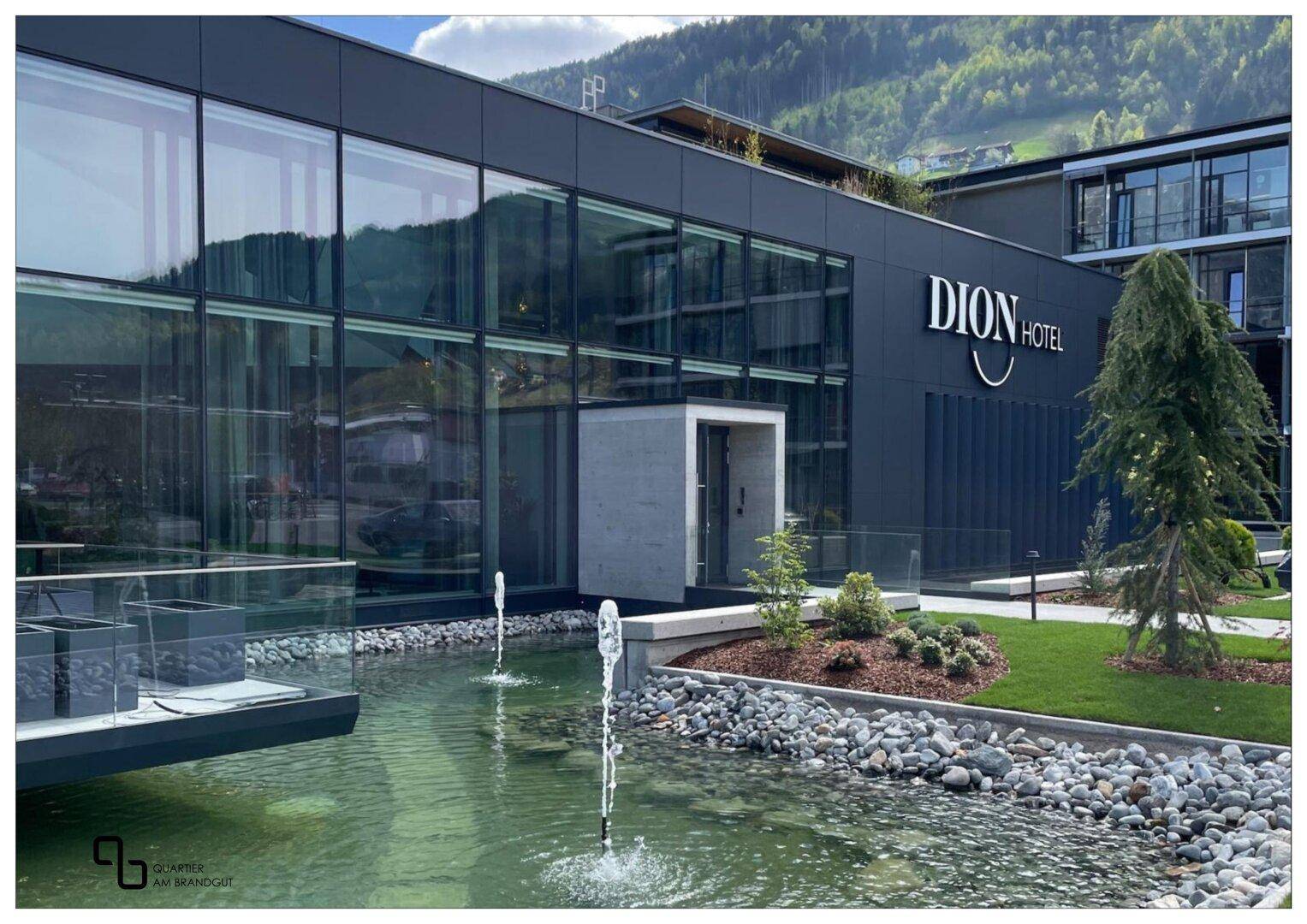 Moderne Hotelfassade mit großen Glasflächen, Wasserspiel und gepflegter Außenanlage vor Bergkulisse.