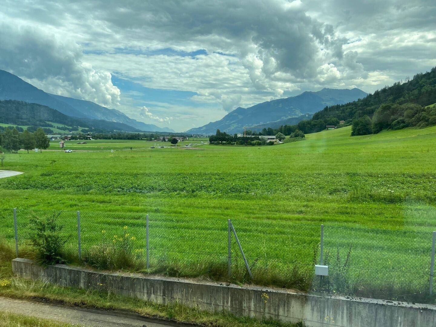 Weitläufige grüne Landschaft mit Feldern und Bergen unter einem bewölkten Himmel, die eine malerische Aussicht bietet.