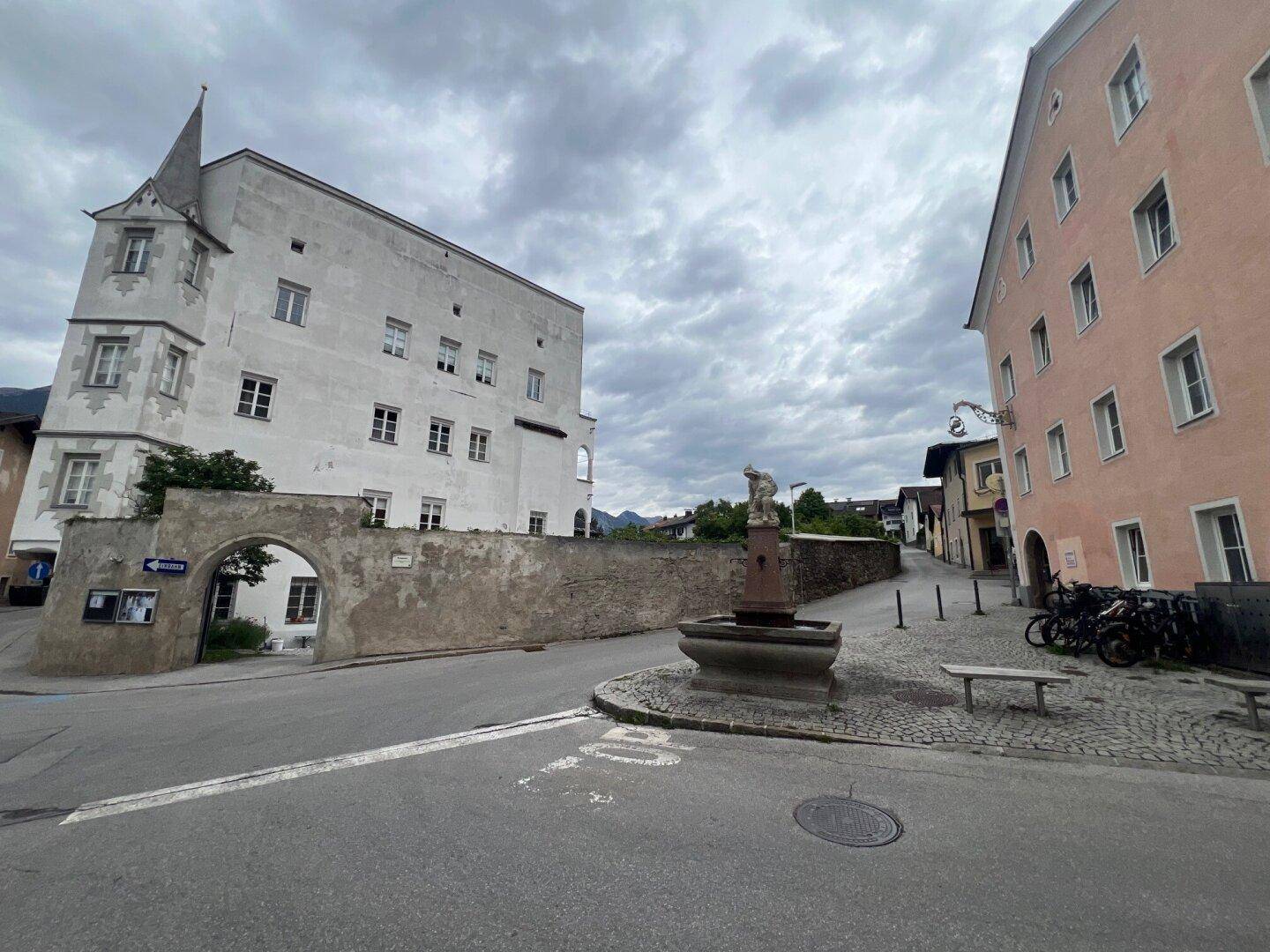 Blick auf eine historische Gasse mit einem Brunnen und zwei alten Gebäuden unter bewölktem Himmel.
