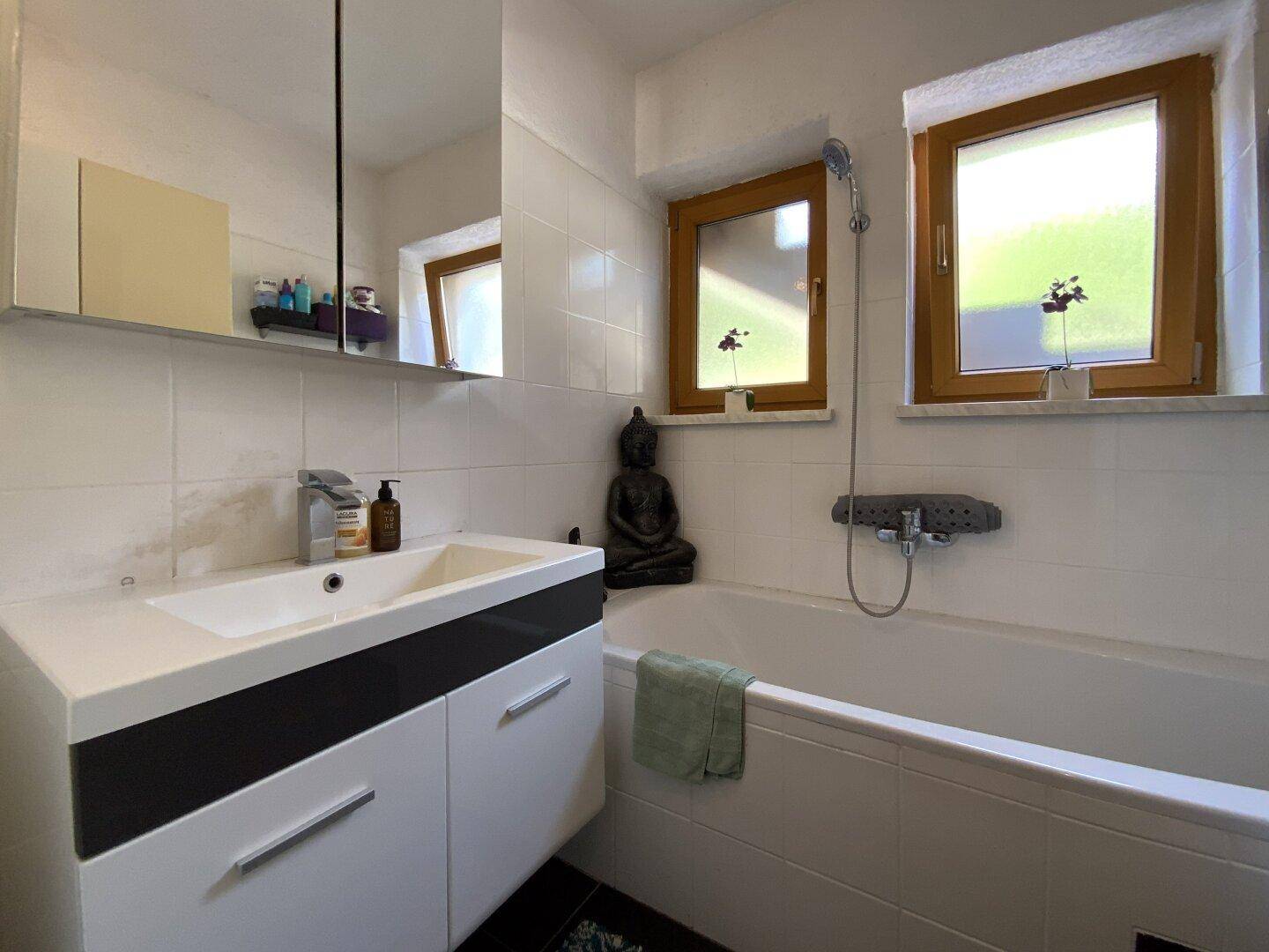 Helles Badezimmer mit Badewanne, Waschtisch und zwei Fenstern für Tageslicht.