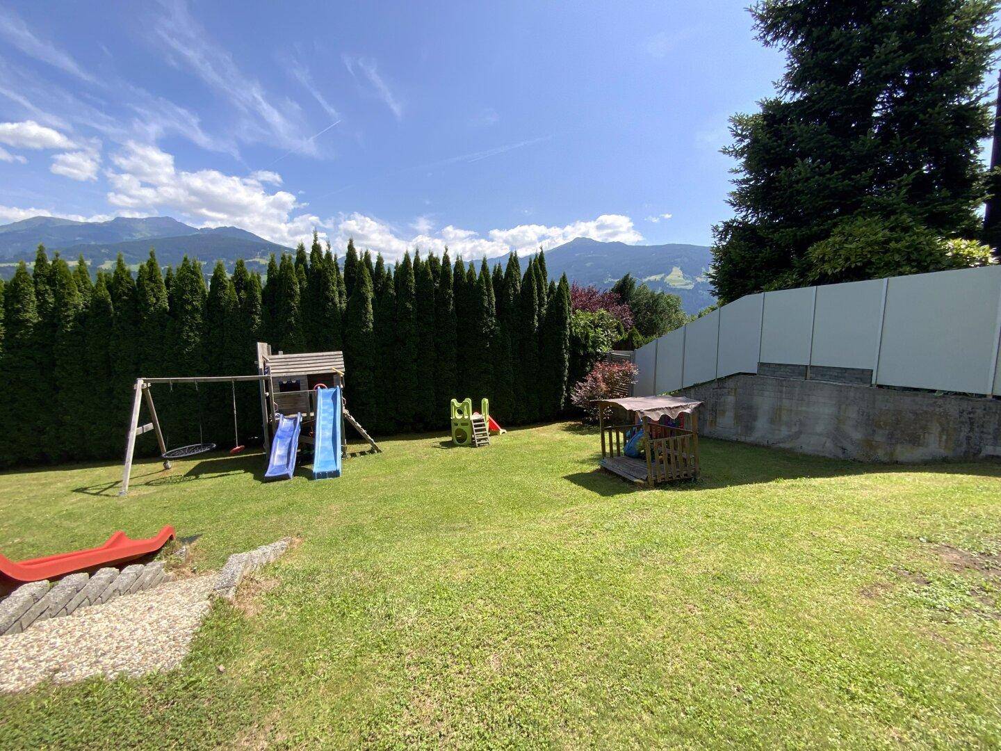 Großer Garten mit Spielplatz, Schaukeln und Rutschen, umgeben von einer hohen Hecke und Bergblick.