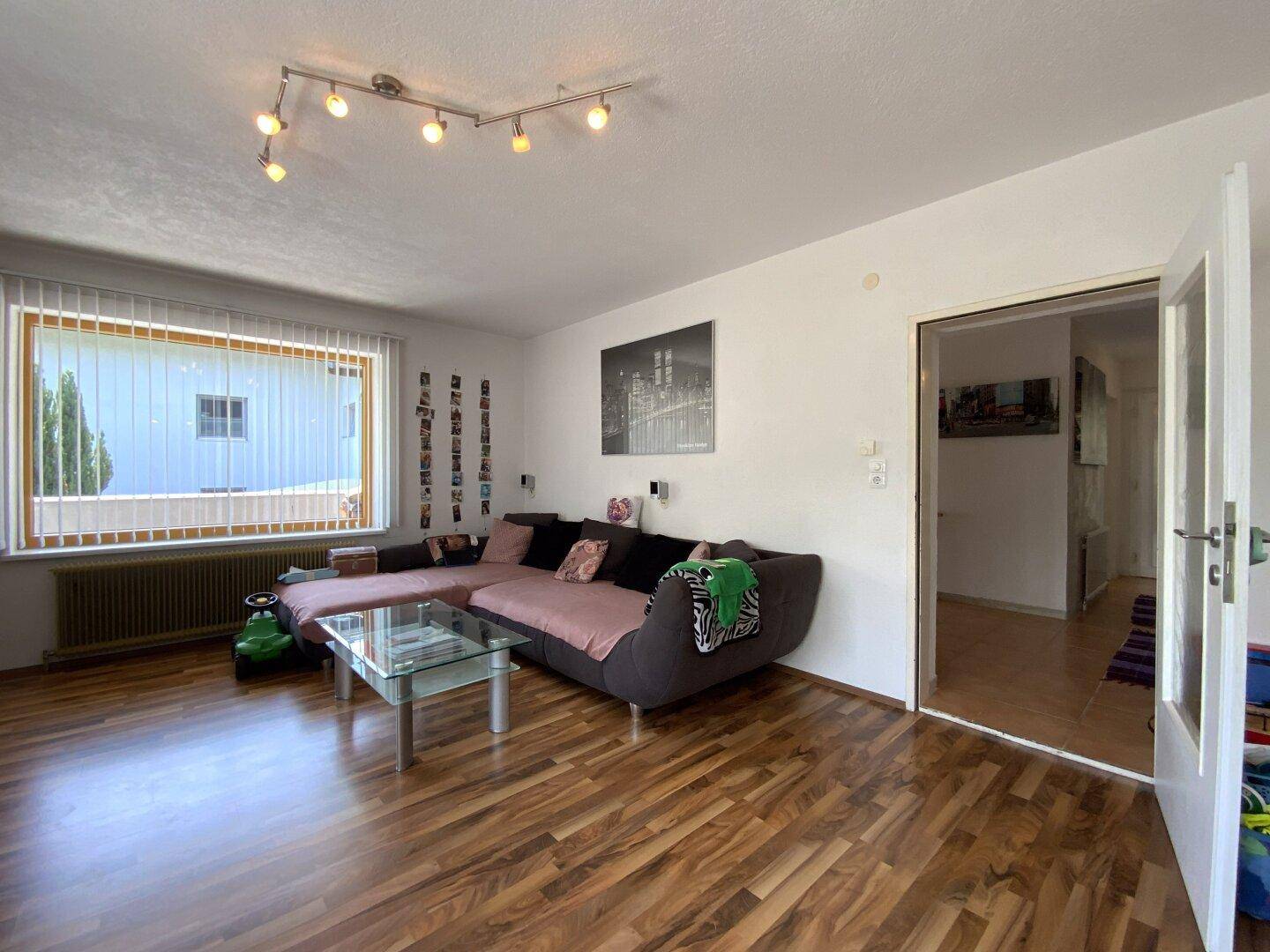 Helles Wohnzimmer mit Laminatboden, großem Sofa und Fenster mit Jalousien.