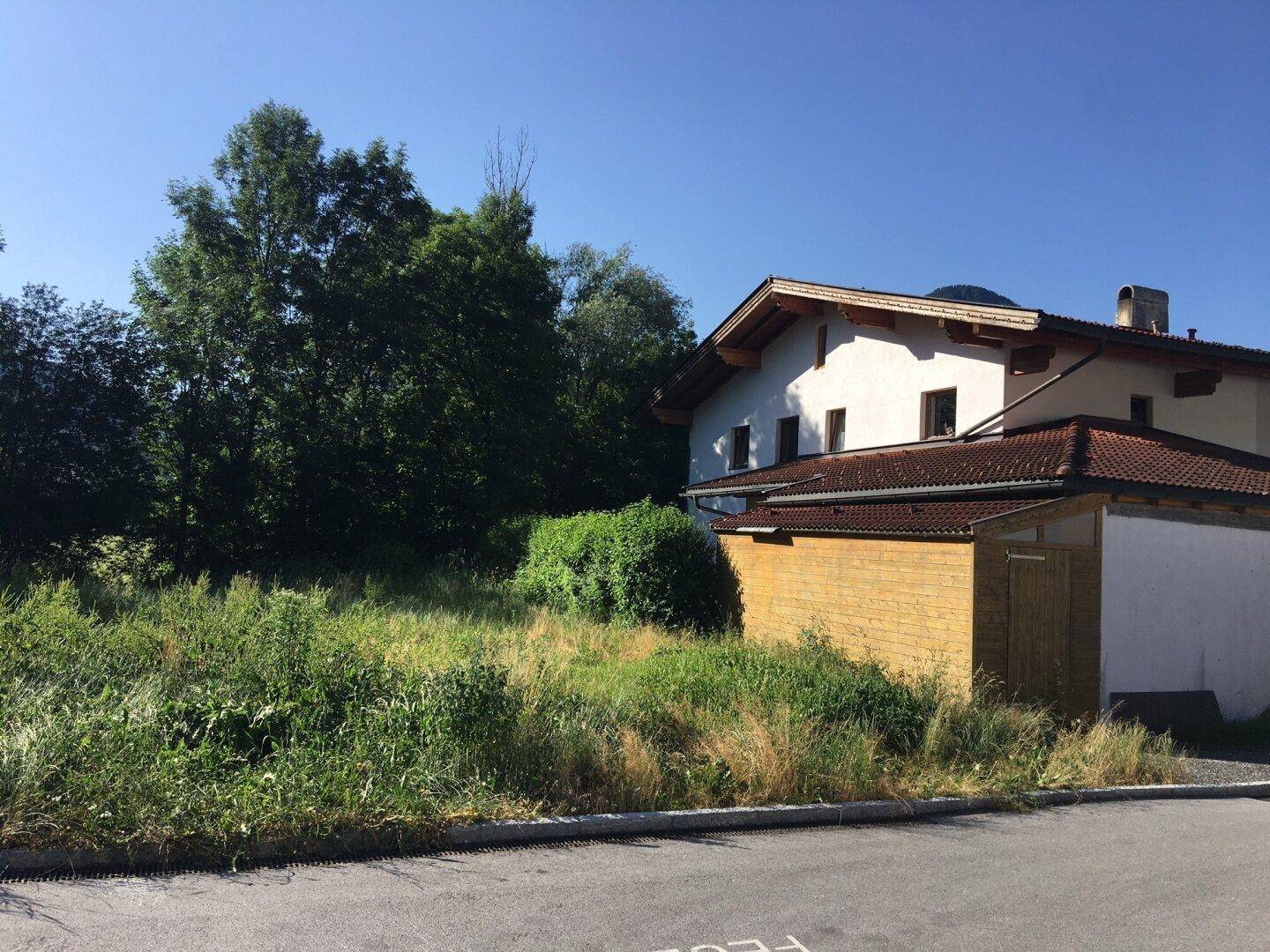 Grünfläche neben einem Wohnhaus mit Holzschuppen und Bergblick unter blauem Himmel.