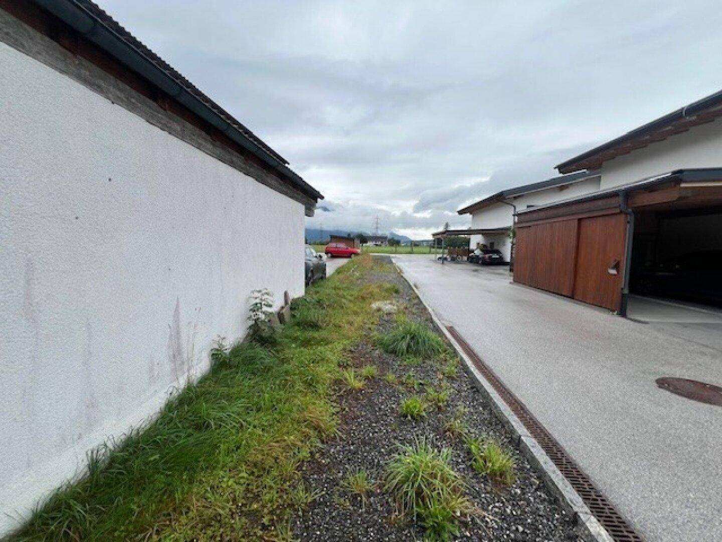 Schmaler Weg zwischen zwei Gebäuden mit Kies und Unkraut unter bewölktem Himmel und Bergsicht.