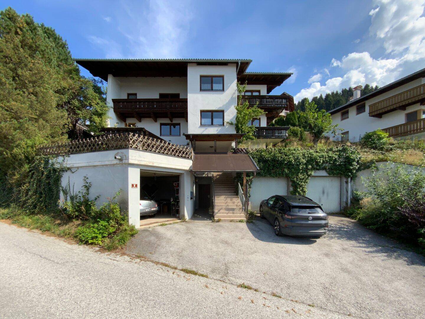 Mehrstöckiges Wohnhaus mit Garagen und Zufahrt, eingebettet in eine grüne Hanglage.