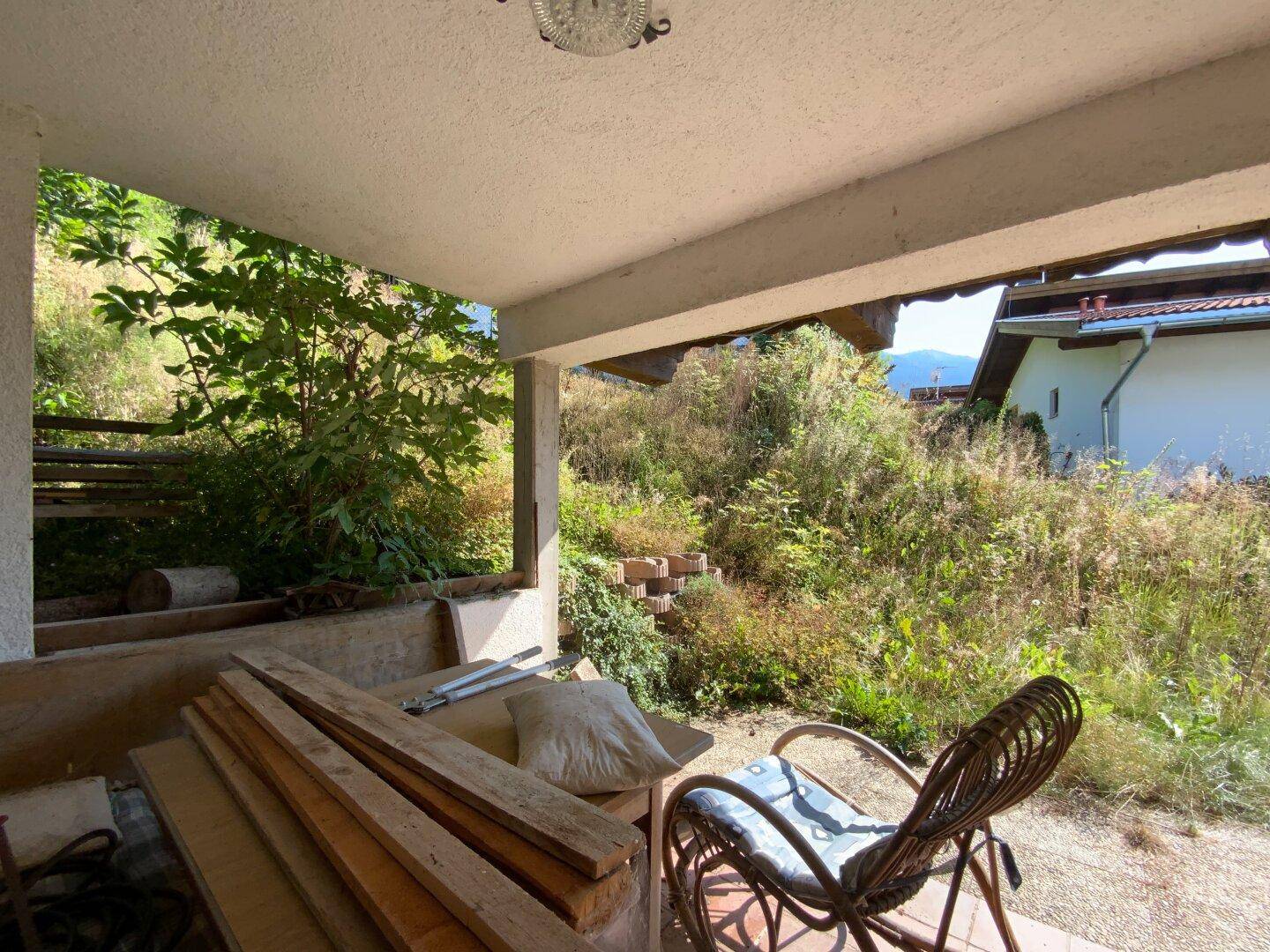 Überdachter Außenbereich mit Holzlatten und einem Korbstuhl, umgeben von viel Grün.