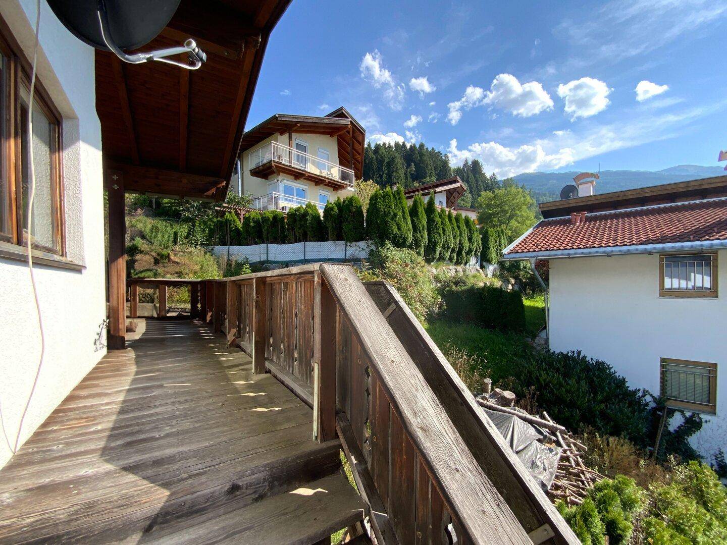 Langer Holzsteg entlang des Gebäudes mit Treppe, umgeben von Bäumen und anderen Häusern.