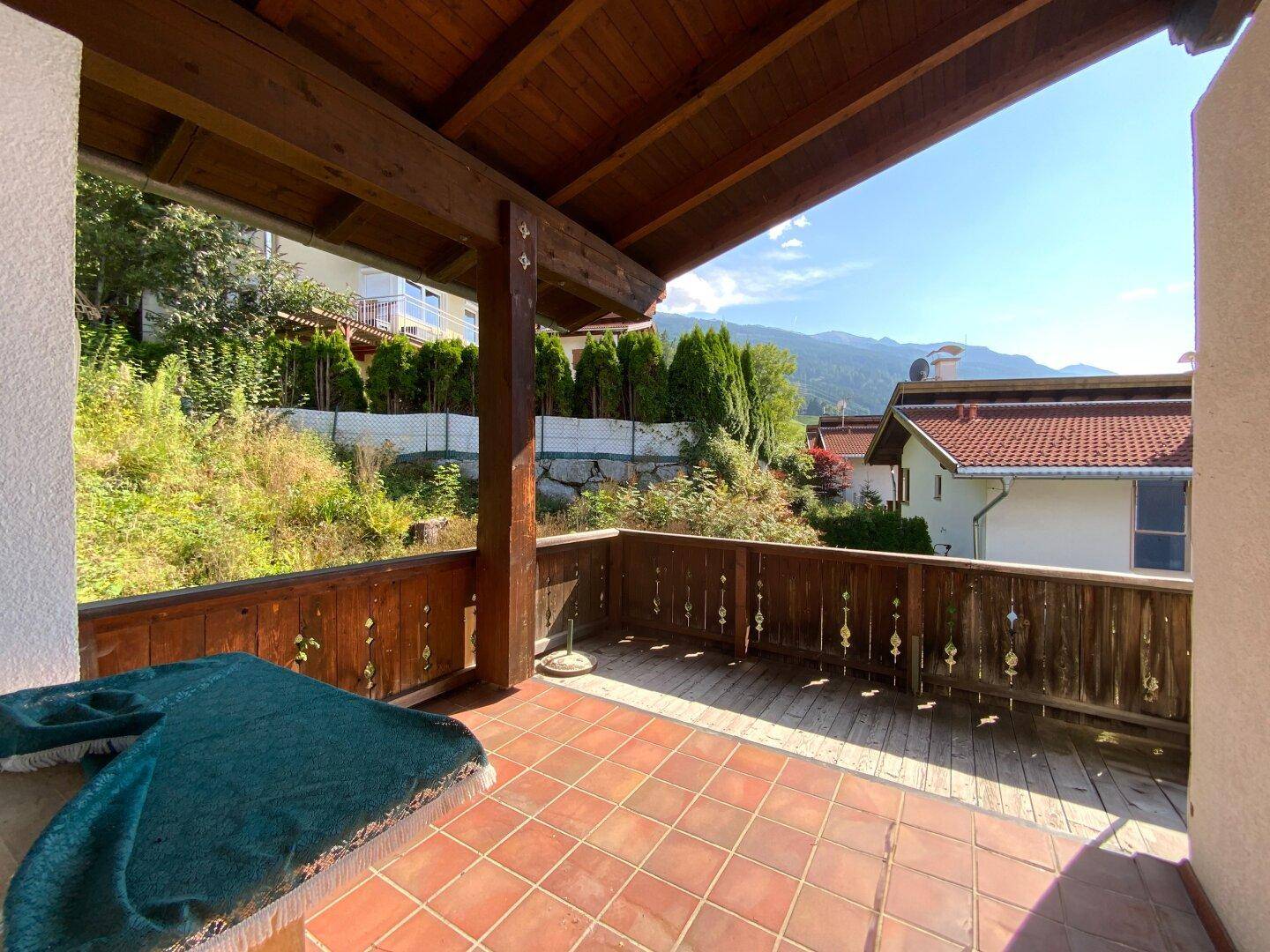 Überdachter Balkon mit Fliesenboden und Holzgeländer, bietet Blick auf den bewachsenen Hang und Berge.