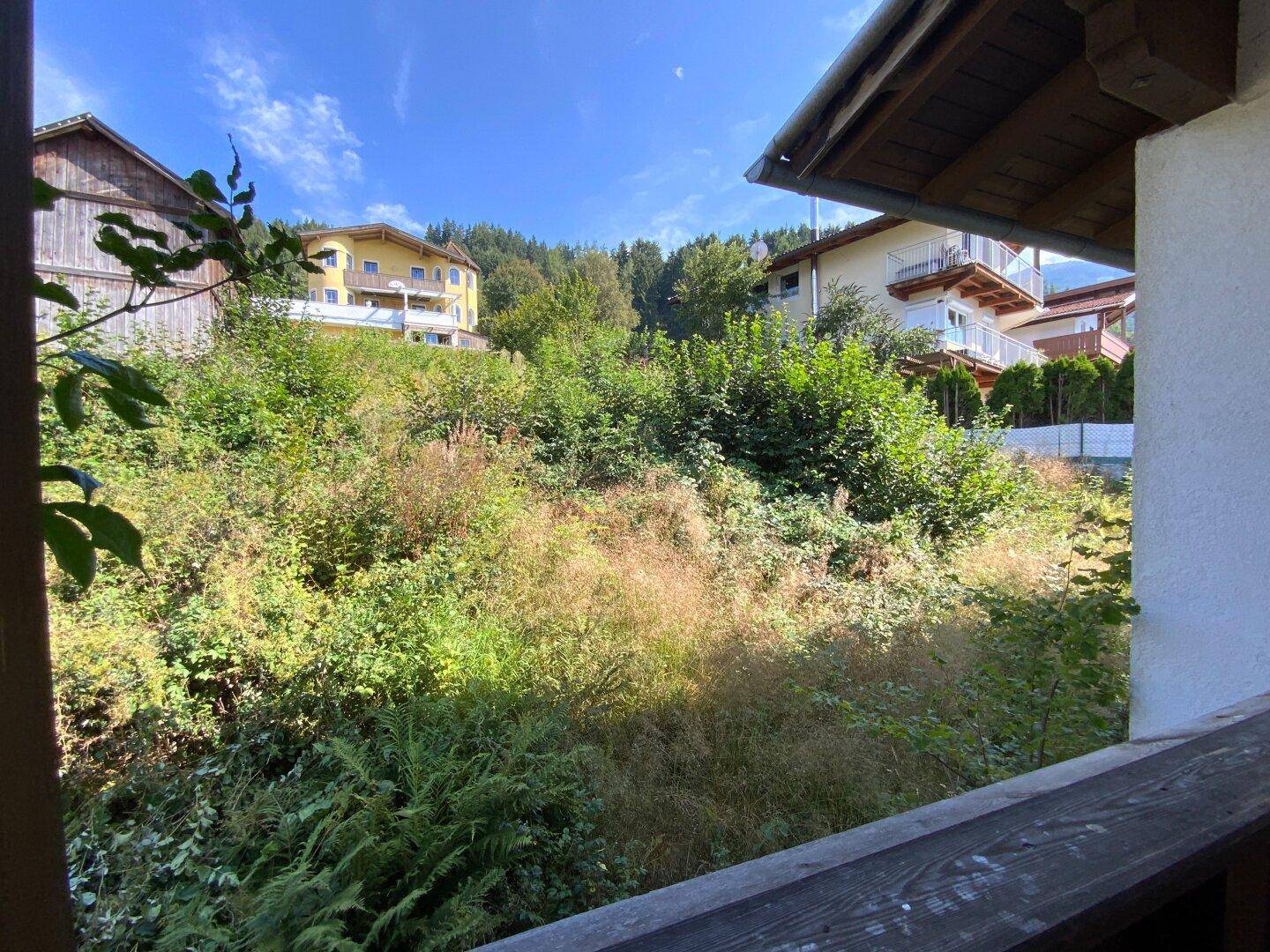Aussicht von einem Balkon auf einen grünen, bewachsenen Hang und benachbarte Gebäude.
