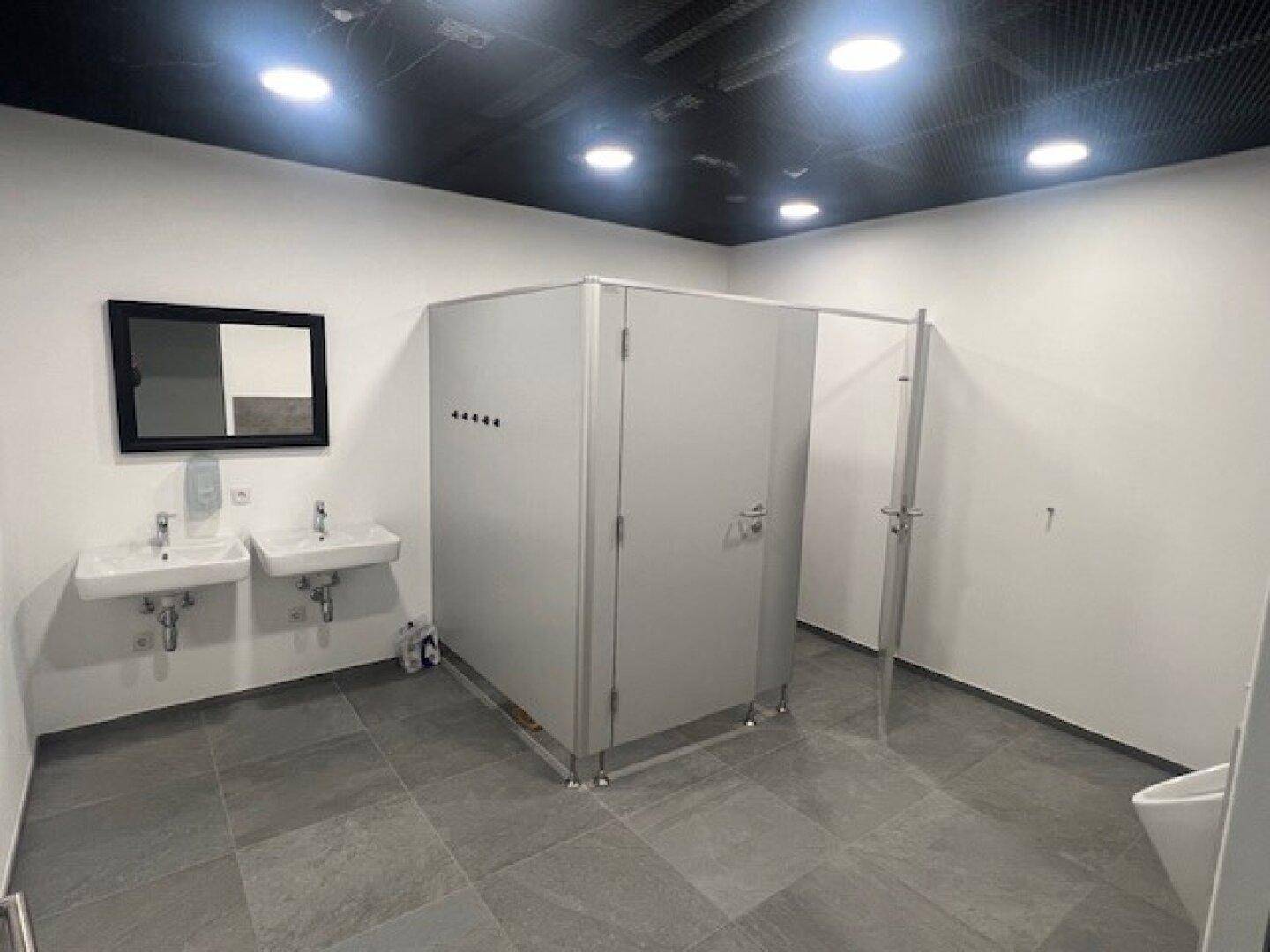 Moderner Sanitärbereich mit zwei Waschbecken, Spiegel und Toilettenkabinen, ausgestattet mit Fliesenboden.