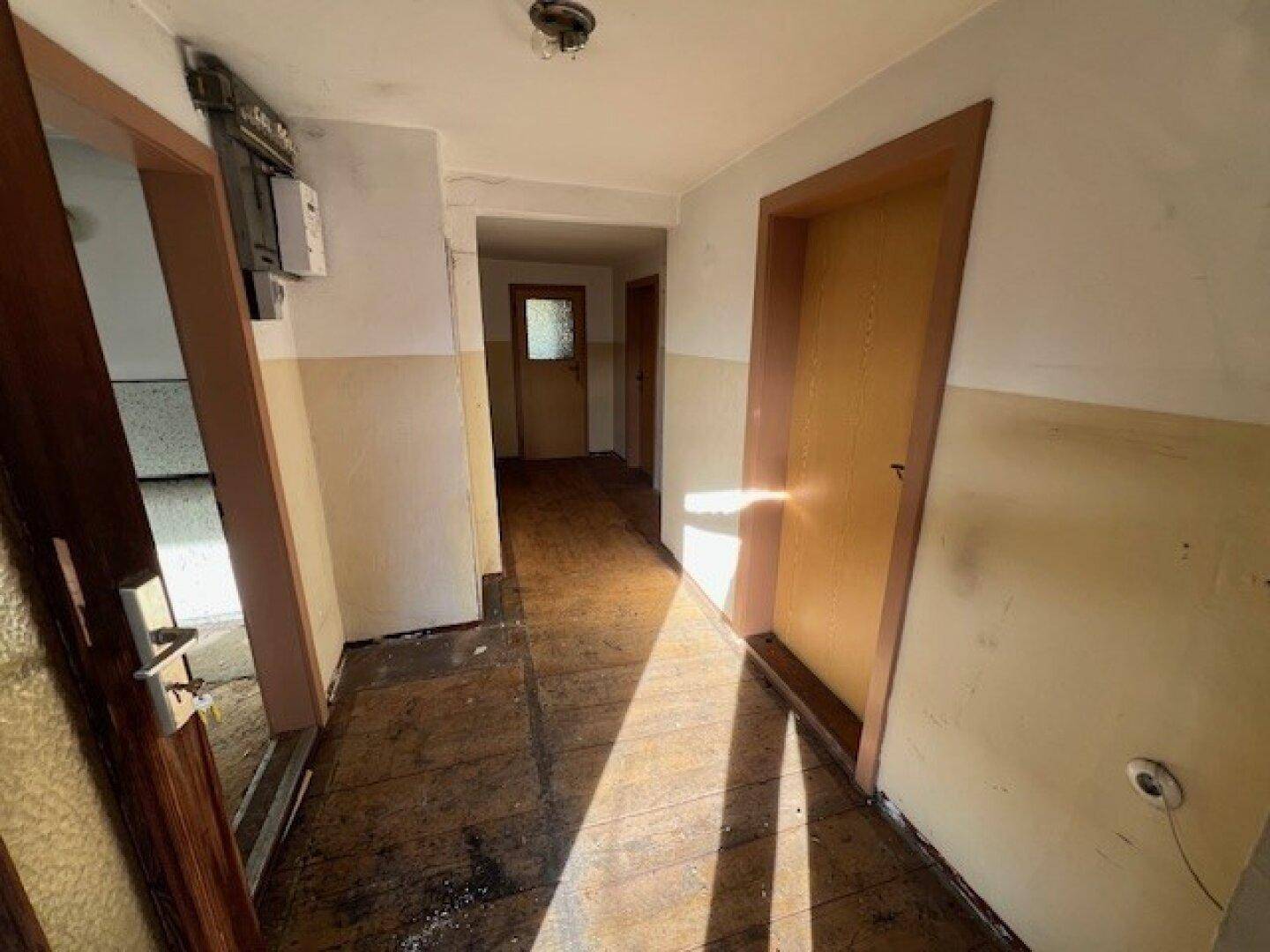 Langer Flur mit abgenutztem Holzfußboden und hellen Wänden, durchflutet von Sonnenlicht.