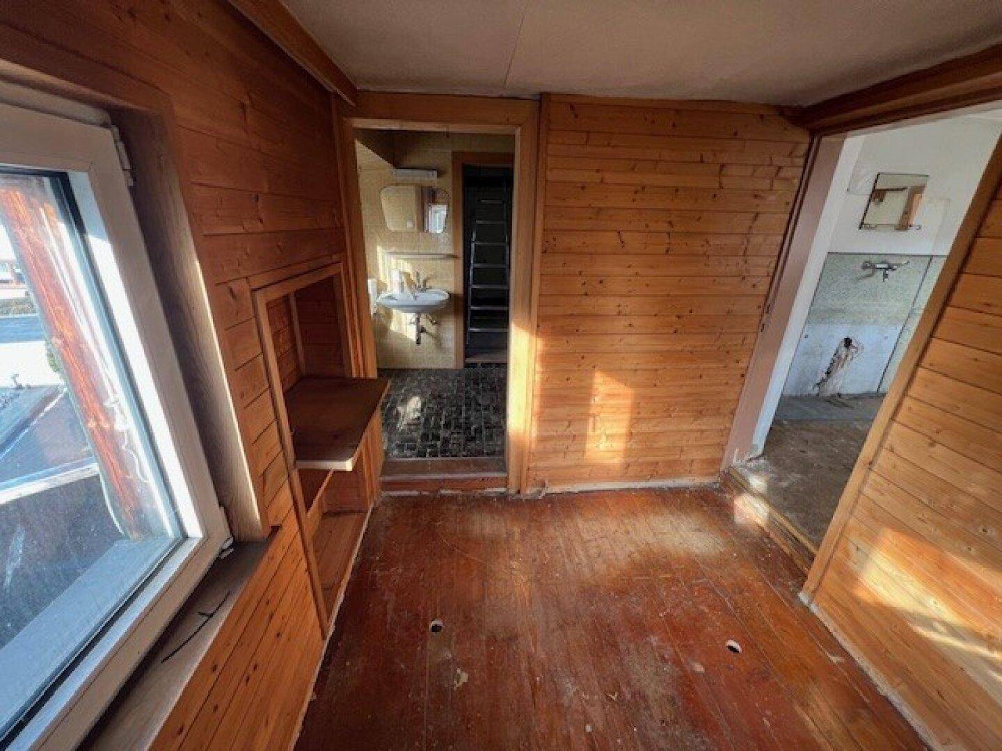 Raum mit Holzvertäfelung und Blick in ein angrenzendes Badezimmer mit Waschbecken.