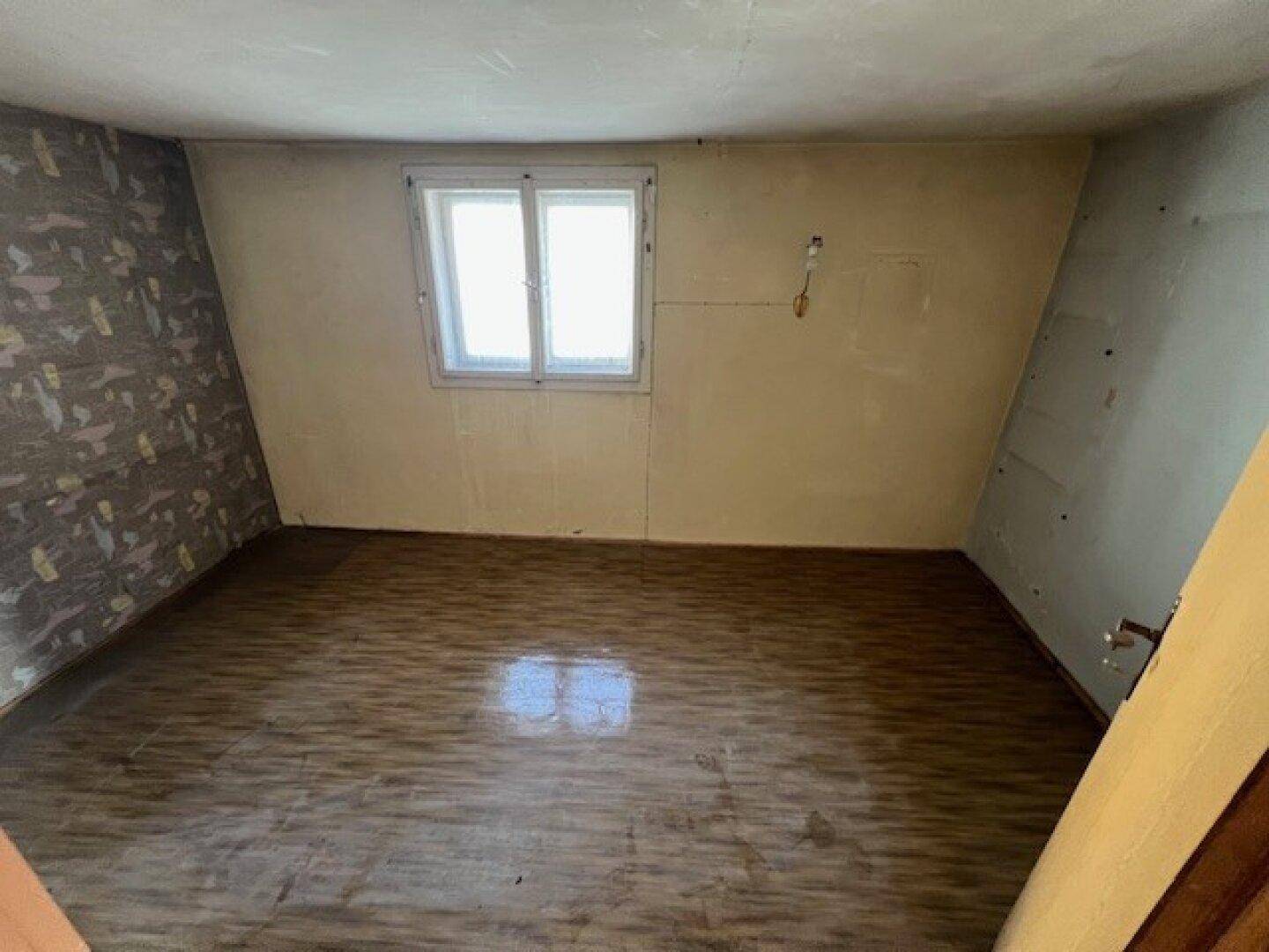 Leeres Zimmer mit Fenster, gemusterter Tapete und abgenutztem Holzfußboden.