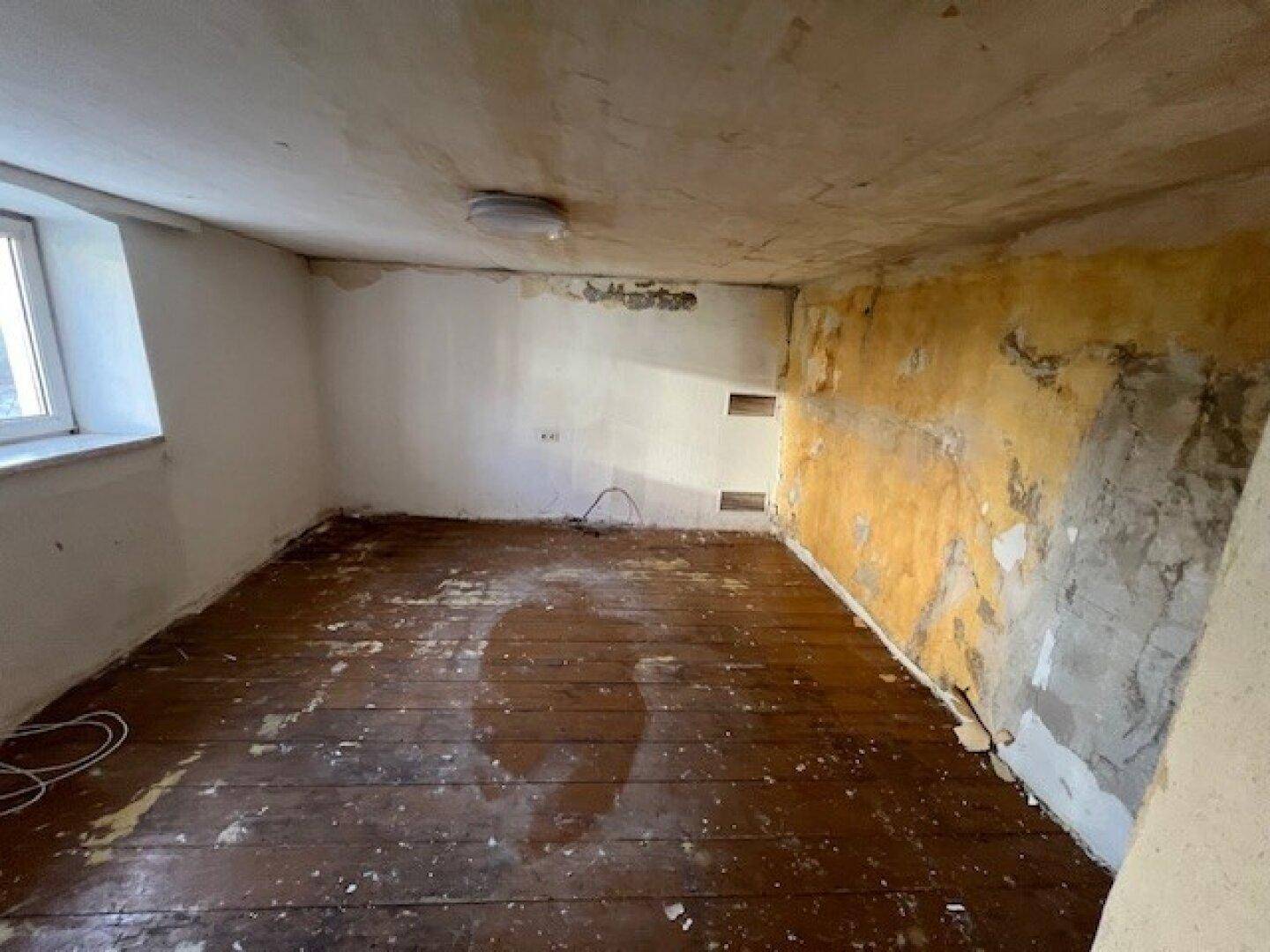Leeres Zimmer mit abgenutztem Holzfußboden und teilweise abgezogener Tapete.