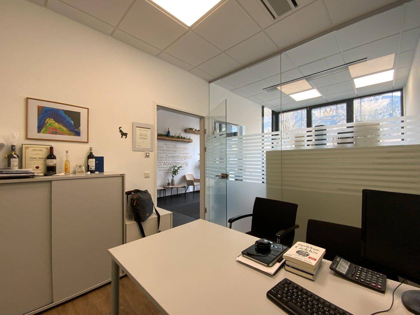 Büro mit Schreibtisch, Computer und Glaswand, die den Raum vom Flur trennt.