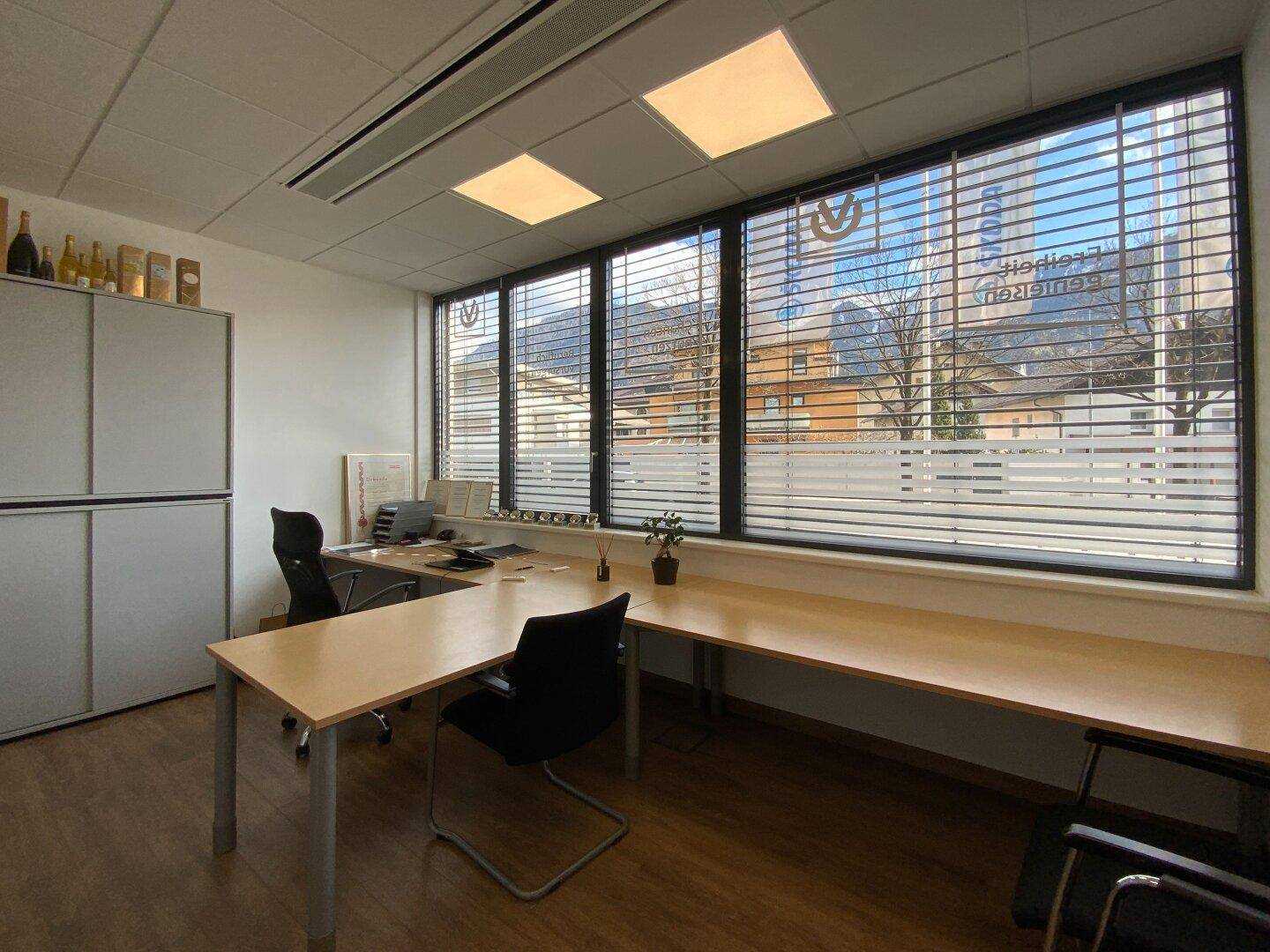 Funktionales Büro mit langen Schreibtischen, Bürostühlen und Jalousien an den Fenstern.