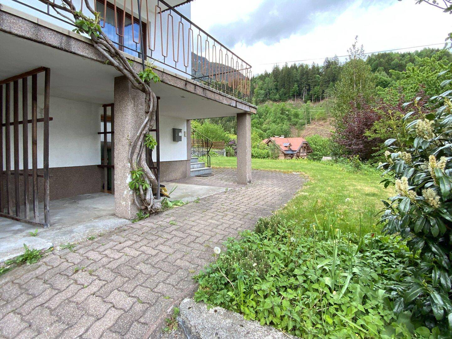 Seitlicher Gartenbereich mit gepflastertem Weg, Rasenfläche und überdachtem Zugang unter dem Balkon.