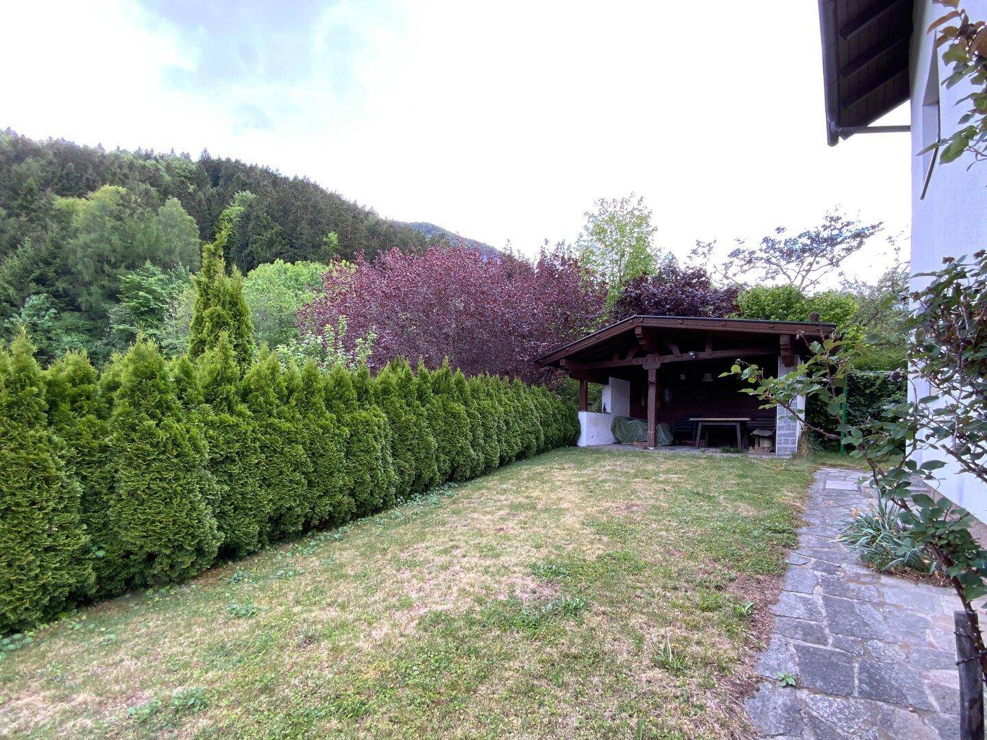 Gepflegter Garten mit hoher Hecke, Rasenfläche und einer rustikalen Holzpergola im hinteren Bereich.