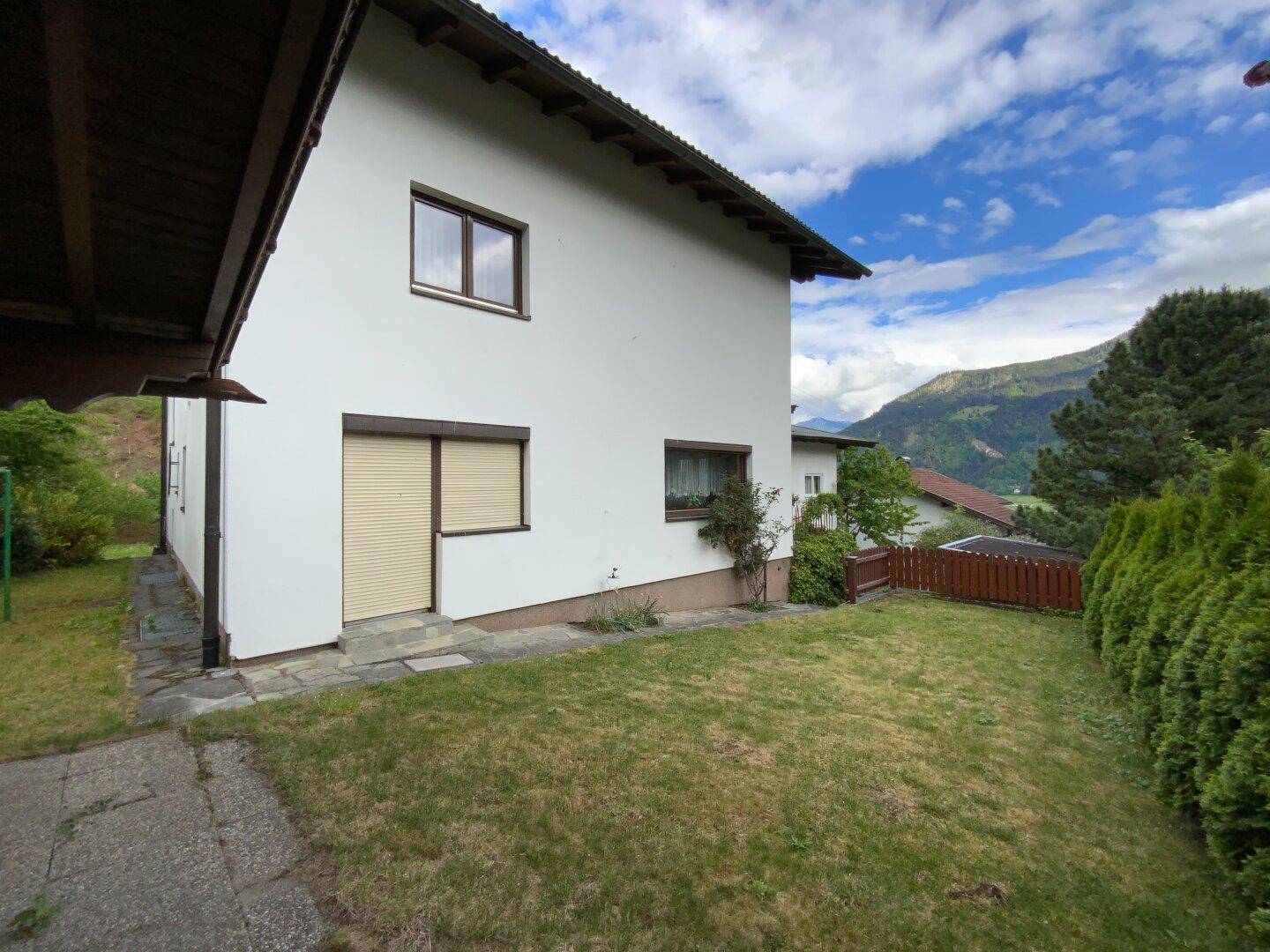 Seitenansicht des Hauses mit weißer Fassade, Rollläden an den Fenstern und einem kleinen Rasenstück.