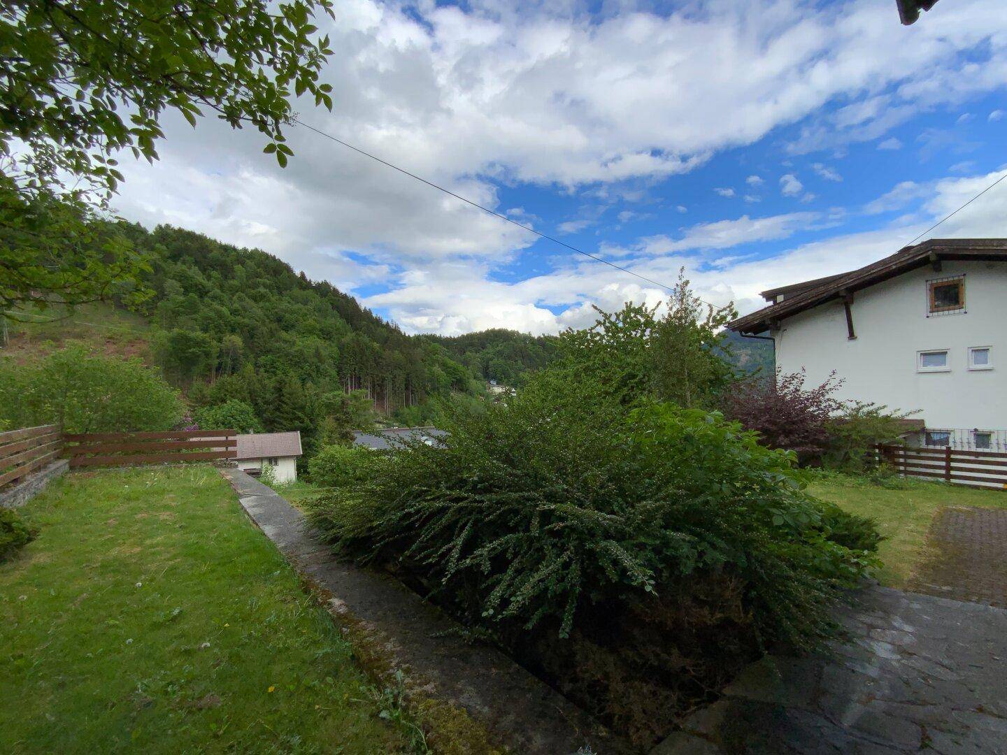 Aussicht von erhöhter Position auf einen grünen Hang, Büsche und ein entferntes Haus unter blauem Himmel.