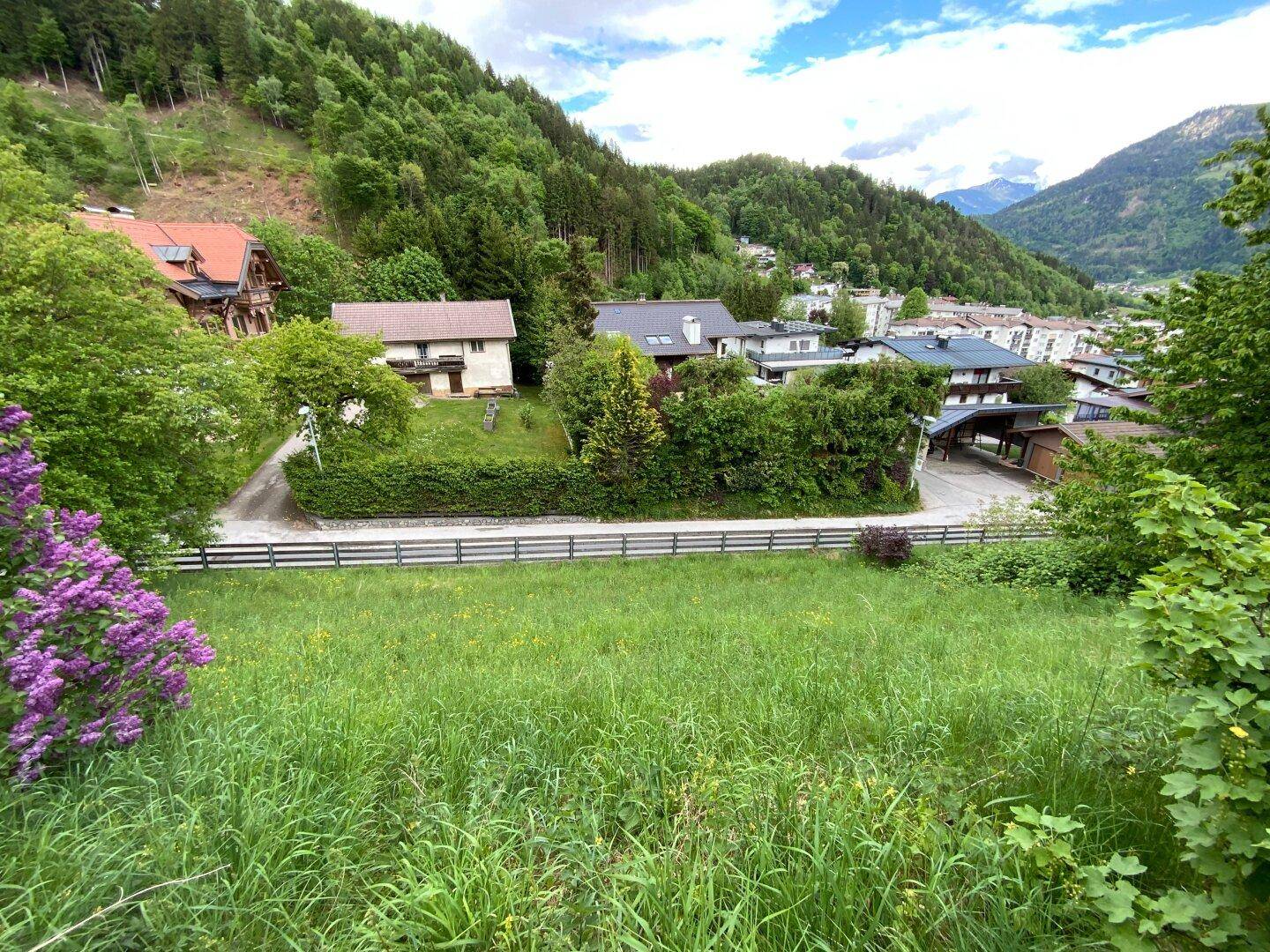 Panoramablick über das Tal mit grünen Wiesen, umliegenden Häusern und majestätischen Bergen im Hintergrund.