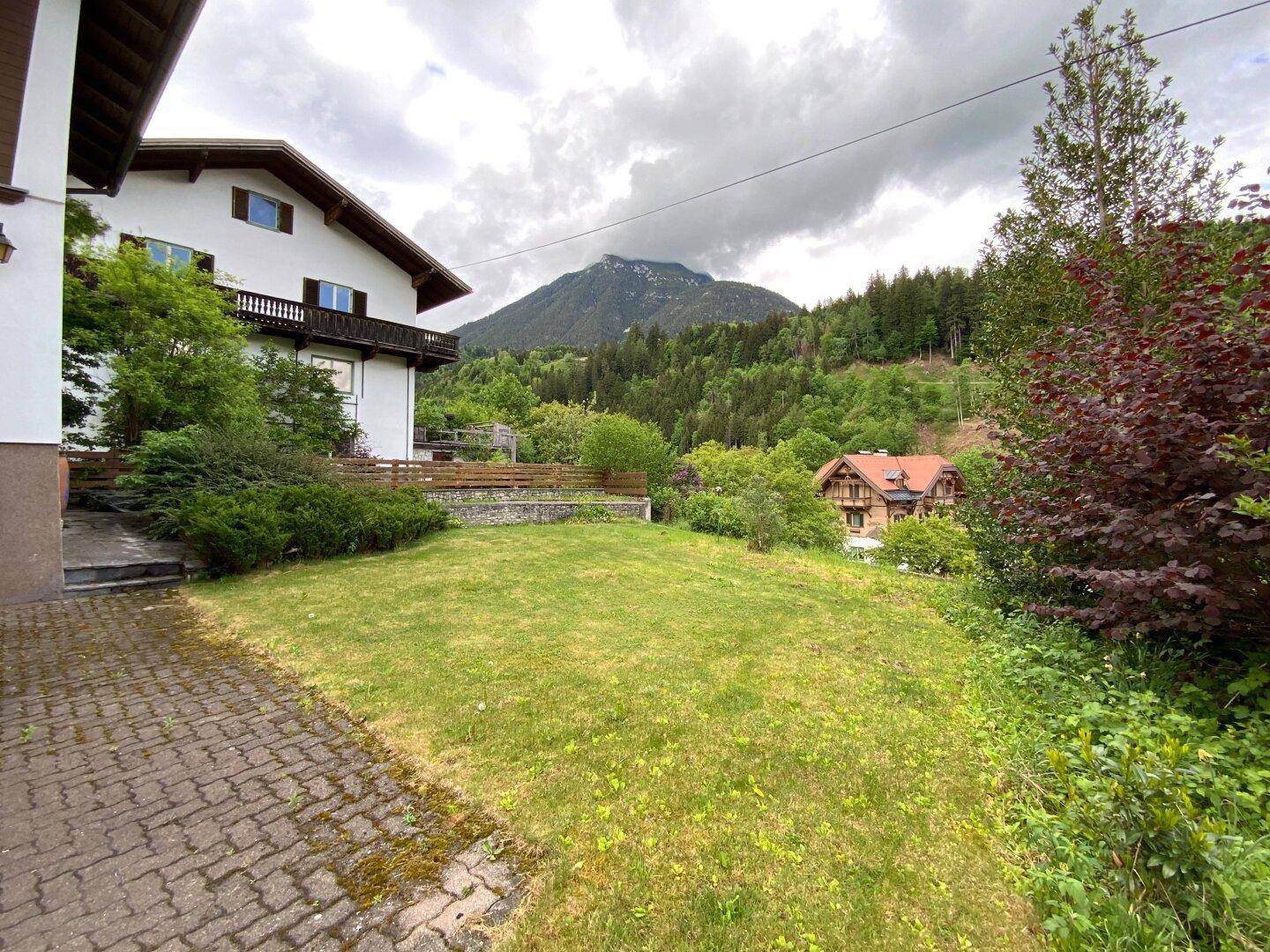 Gartenbereich mit gepflastertem Weg, einem Haus im Hintergrund und einem beeindruckenden Bergblick.
