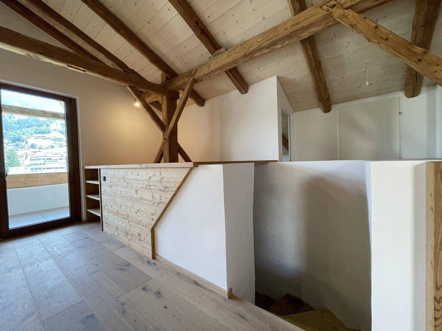 Offener Wohnbereich mit sichtbaren Holzbalken, Holzboden und Zugang zu einem weiteren Bereich.