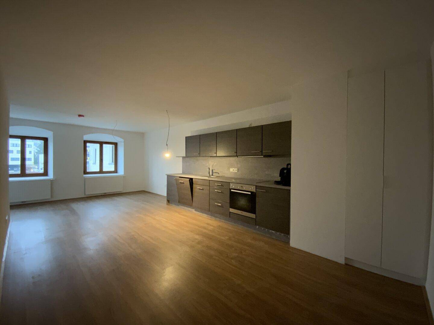 Geräumiger Wohn-Essbereich mit dunkler Einbauküche, Holzboden und zwei Fenstern.
