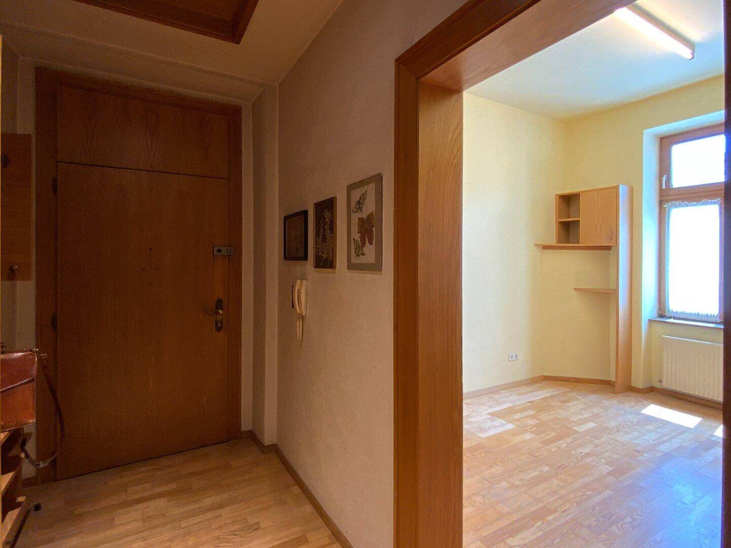 Flur mit Holzboden und Zugang zu einem leeren Zimmer, die Haustür ist sichtbar.