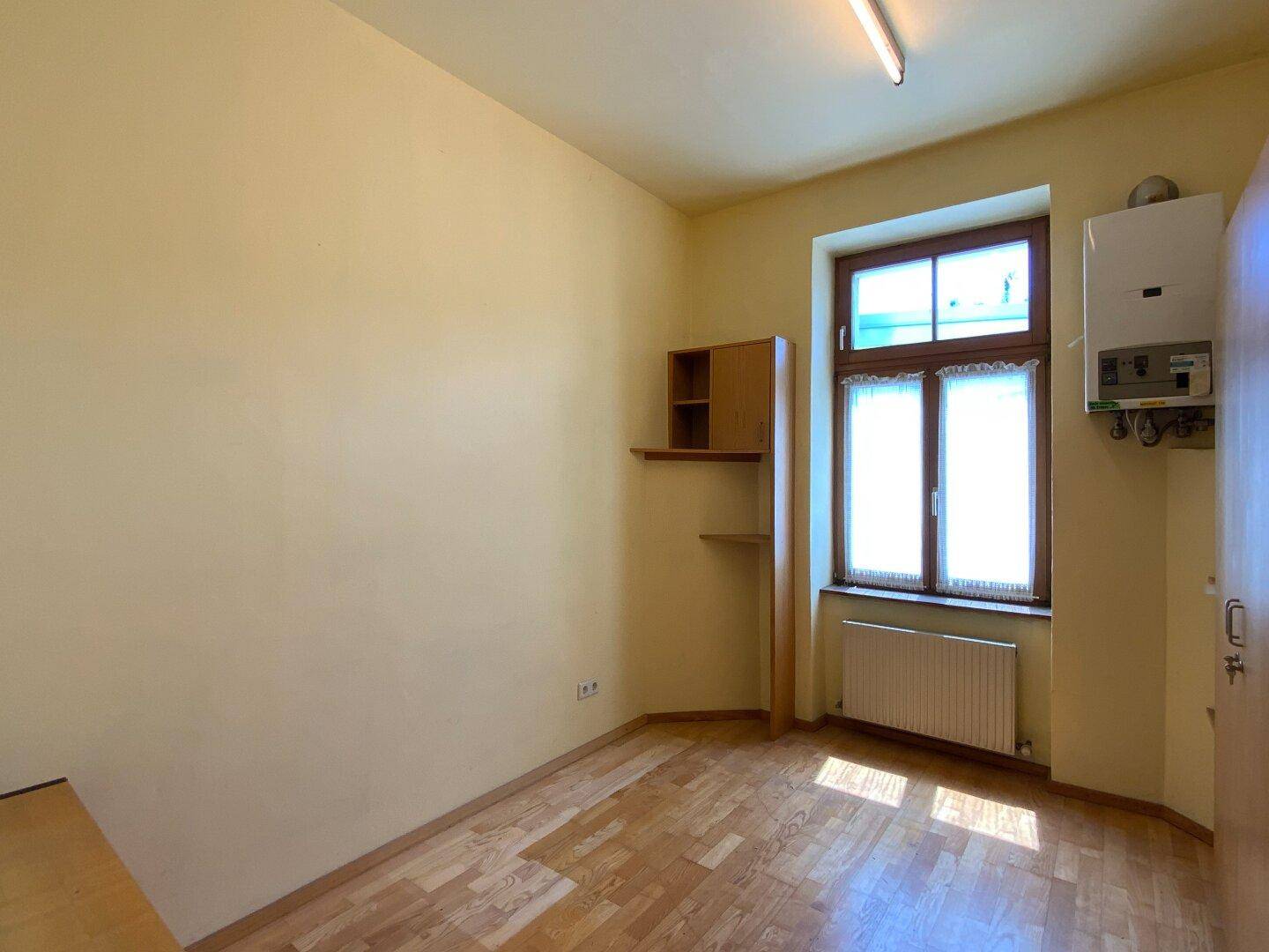 Leeres Zimmer mit Fenster, Heizkörper und an der Wand montiertem Boiler, vielseitig nutzbar.