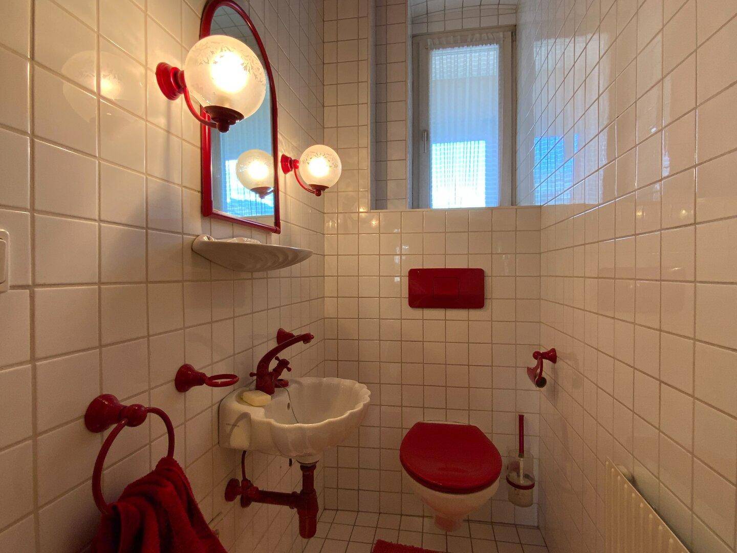 Kleines Gäste-WC mit roten Akzenten, weißen Fliesen und Fenster.