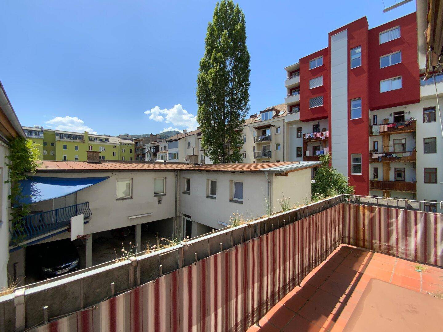 Weitblick von der Loggia auf die umliegenden Gebäude und einen hohen Baum, sonnige Aussicht.