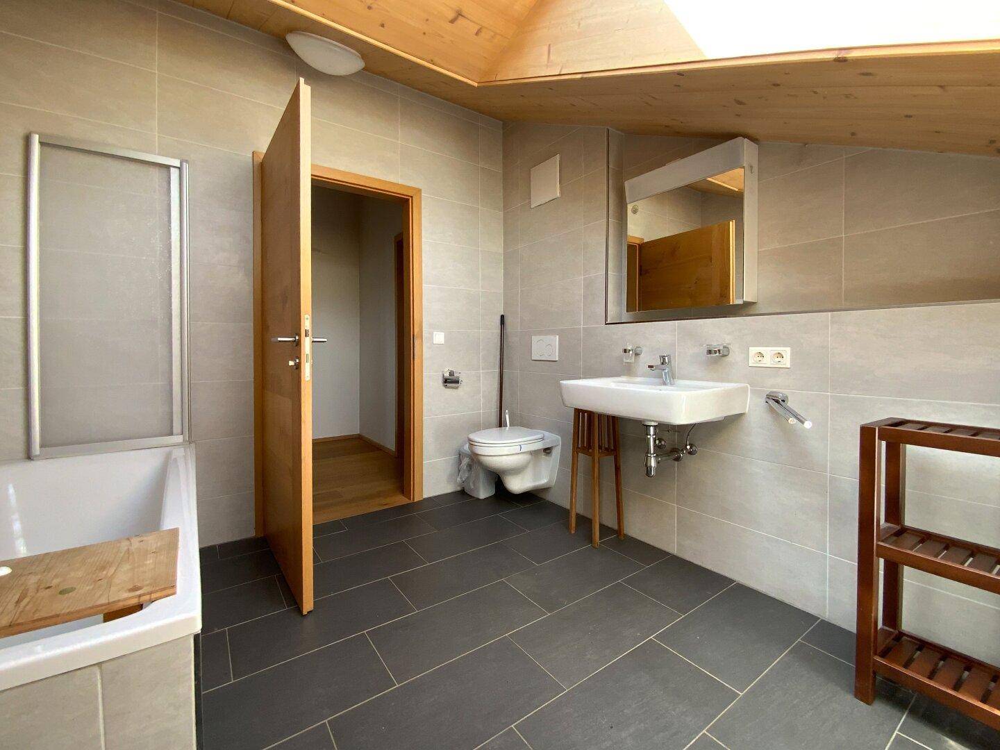 Helles Badezimmer mit grauen Fliesen, Waschbecken, Toilette und Duschbereich.