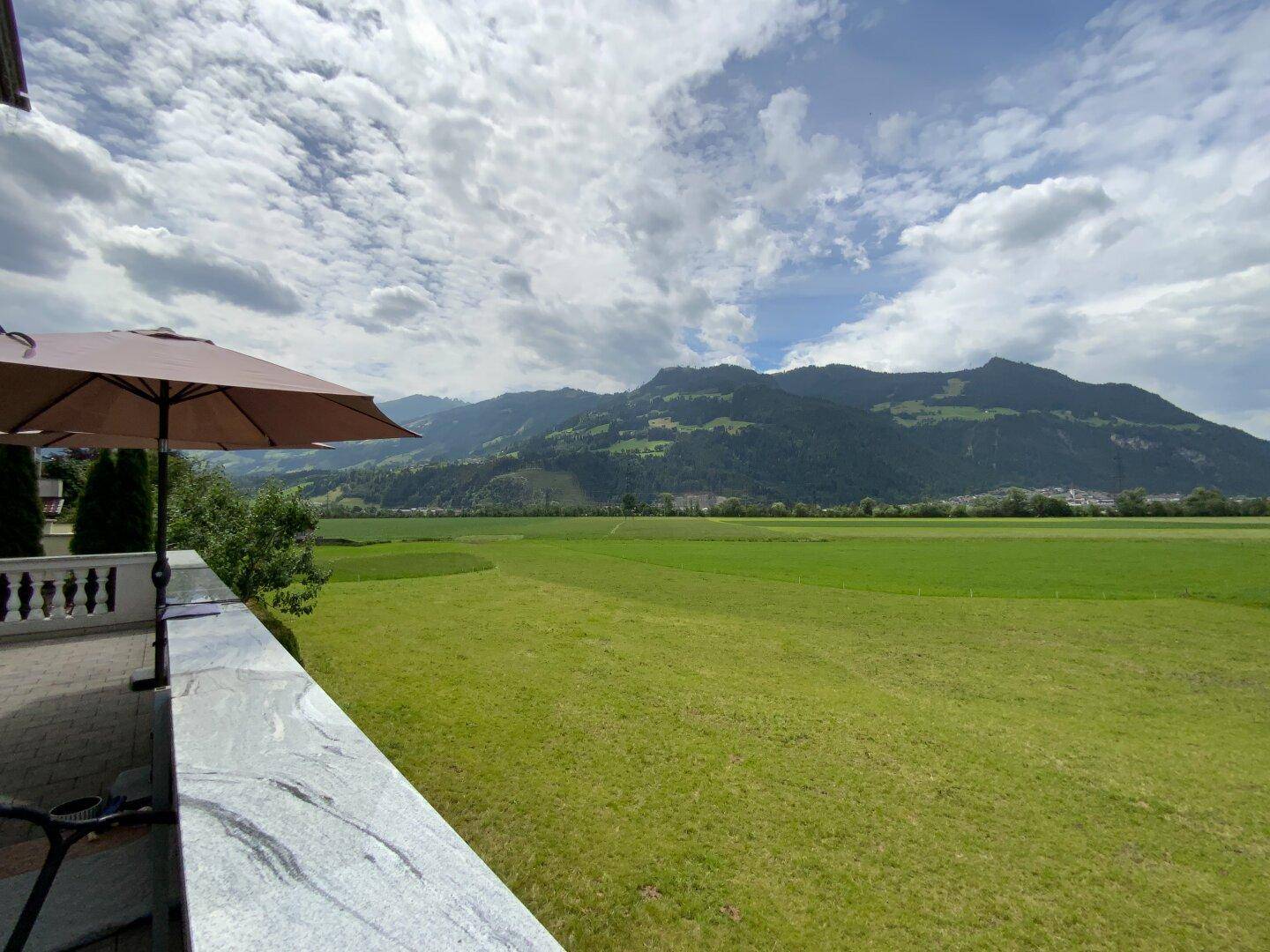 Panoramablick von der Terrasse auf die weiten grünen Felder und die beeindruckende Bergkulisse.