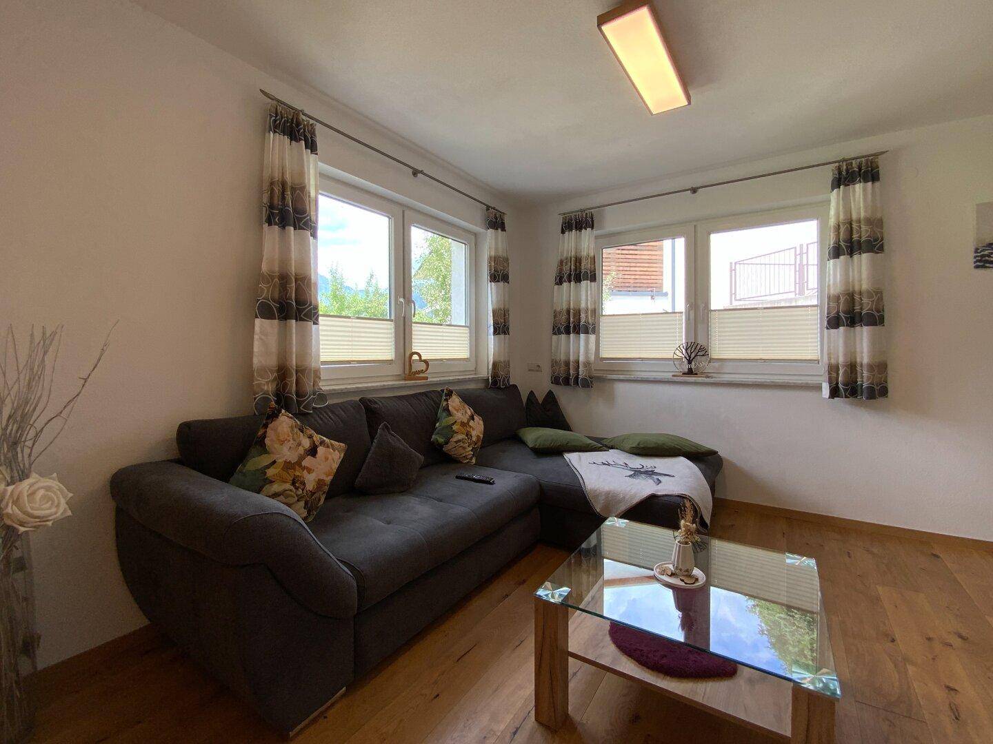 Gemütliches Wohnzimmer mit Ecksofa, Holzboden und mehreren Fenstern für viel Tageslicht.