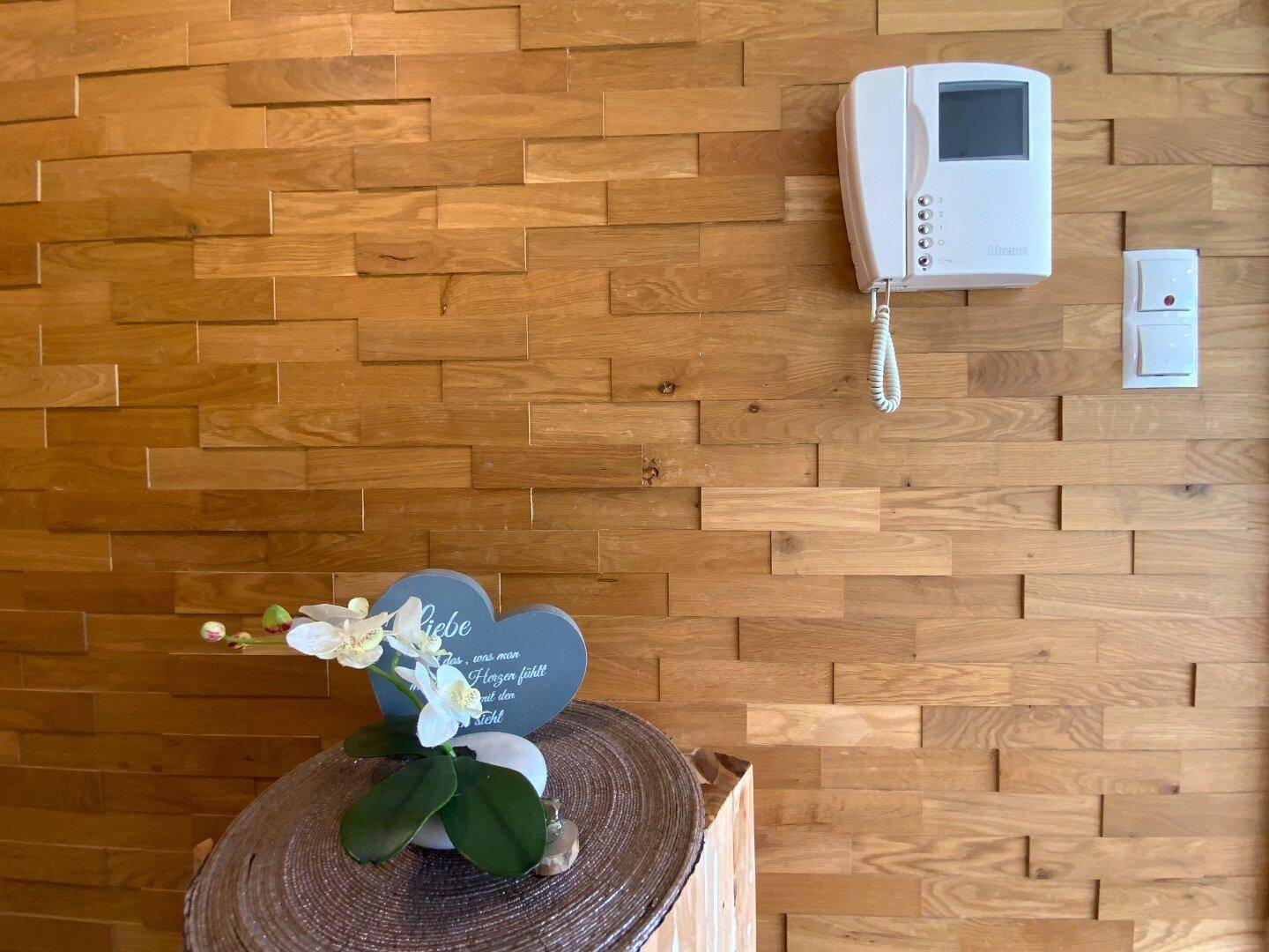 Wandverkleidung aus Holz mit Gegensprechanlage und kleiner Dekoration im Eingangsbereich.
