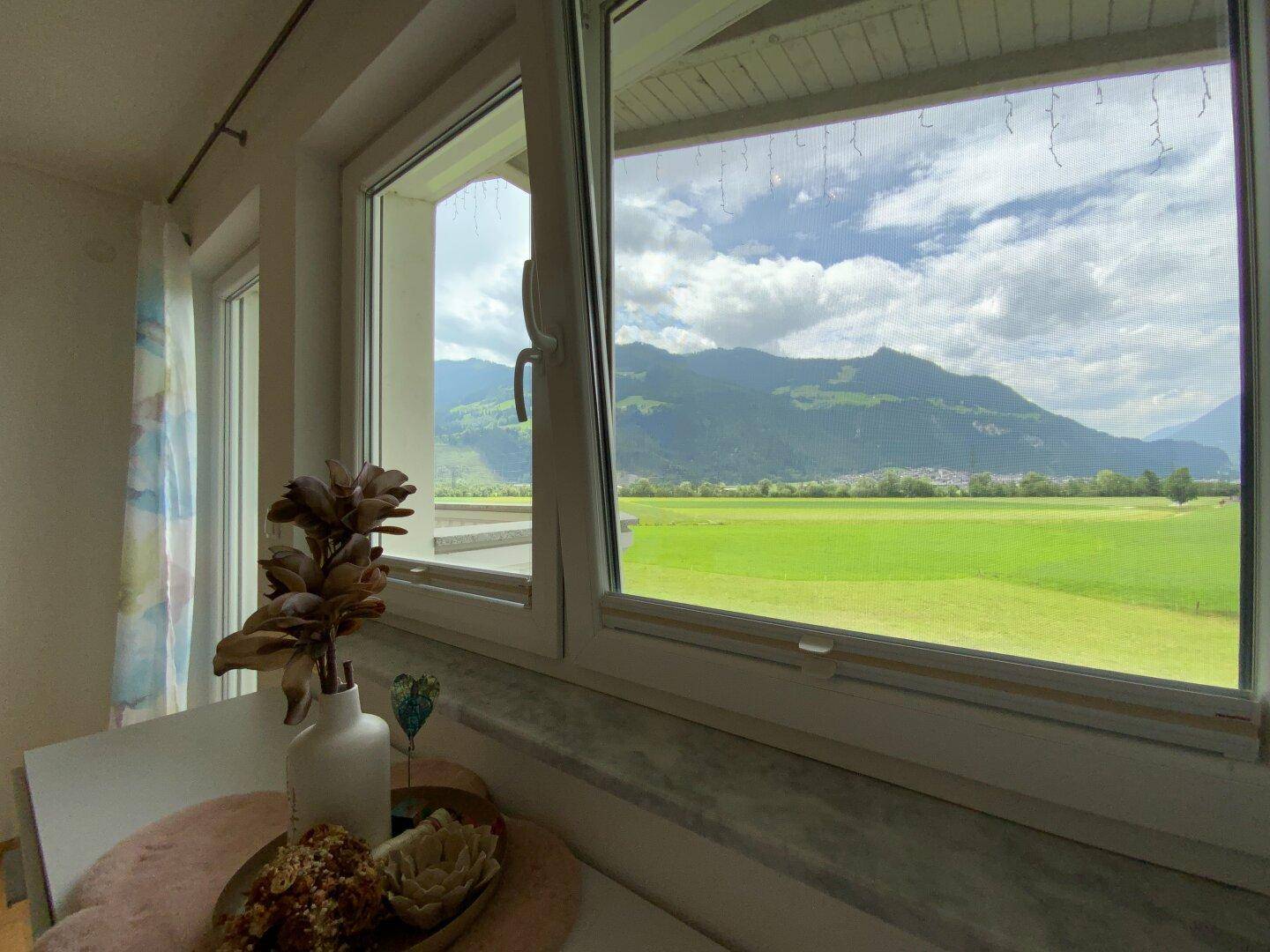 Heller Ausblick aus einem Fenster auf grüne Felder und majestätische Berge.