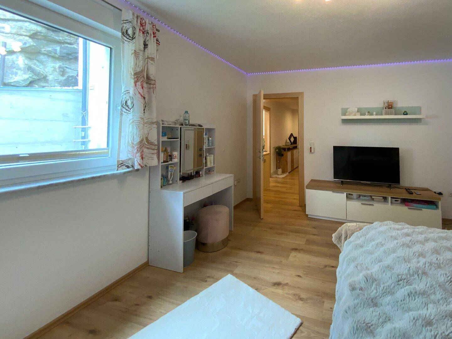 Helles Schlafzimmer mit Schminktisch, Fernseher und Fenster, das eine angenehme Atmosphäre schafft.