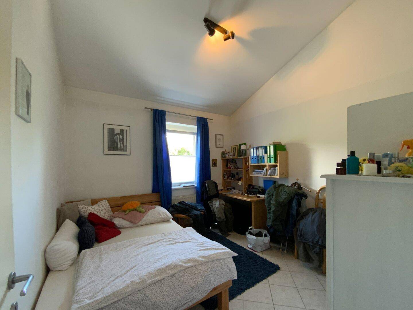 Gemütliches Schlafzimmer mit Einzelbett, Schreibtisch und Dachschräge, hell durch das Fenster.