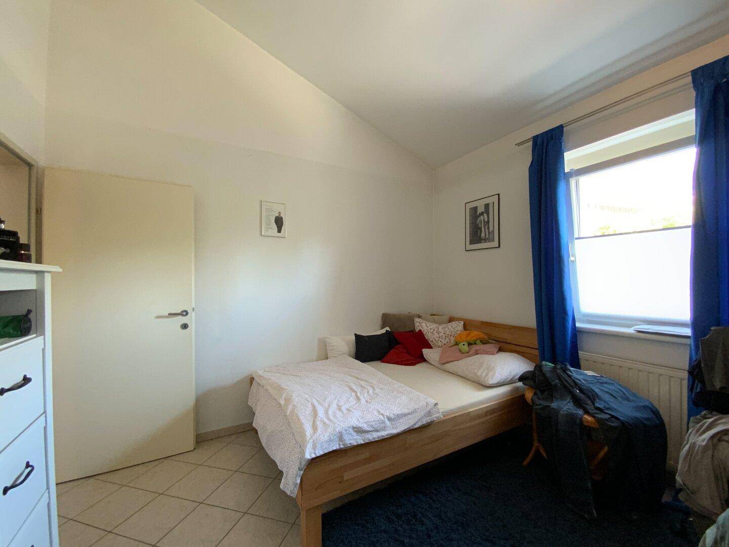 Helles Schlafzimmer mit Einzelbett, Fliesenboden und Fenster mit blauen Vorhängen.